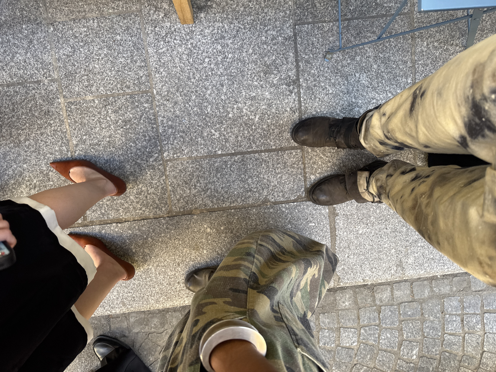 The image shows a view from above of a group of three people standing on a gray stone surface. One person is wearing brown flat shoes with a black and white skirt or dress. Another person is wearing olive-green camouflage pants and dark boots. The third person is wearing pants that are primarily beige with dark brown streaks and dark boots. Part of a metal table leg is visible in the upper right. Part of a wooden table leg is visible in the upper left. The exact location in Paris, France, cannot be determined from the image alone.