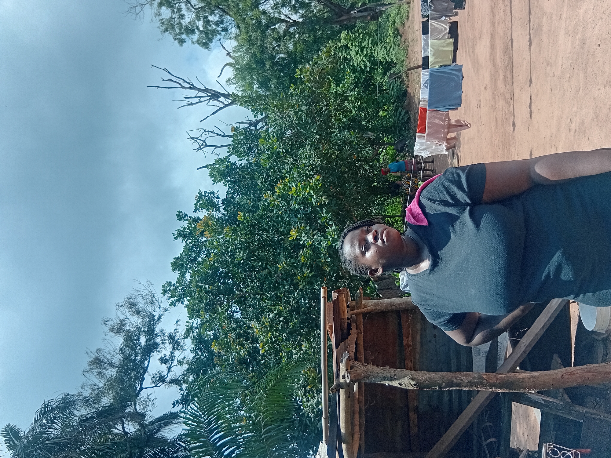 In Ejule, Nigeria, a young Black woman stands in the foreground, her body angled to the right and her head turned to look up and to the right. She wears a black t-shirt with a pink towel draped over her left shoulder. To her left is a rustic wooden structure made of branches and corrugated metal, with a large white bucket at its base. Behind the woman, a clothesline stretches across the frame, laden with an assortment of laundry in various colors, including orange, pink, blue, and white. In the mid-ground, several people are visible near a dense cluster of trees and foliage. The ground is a reddish-brown dirt. The sky above is overcast with gray clouds.