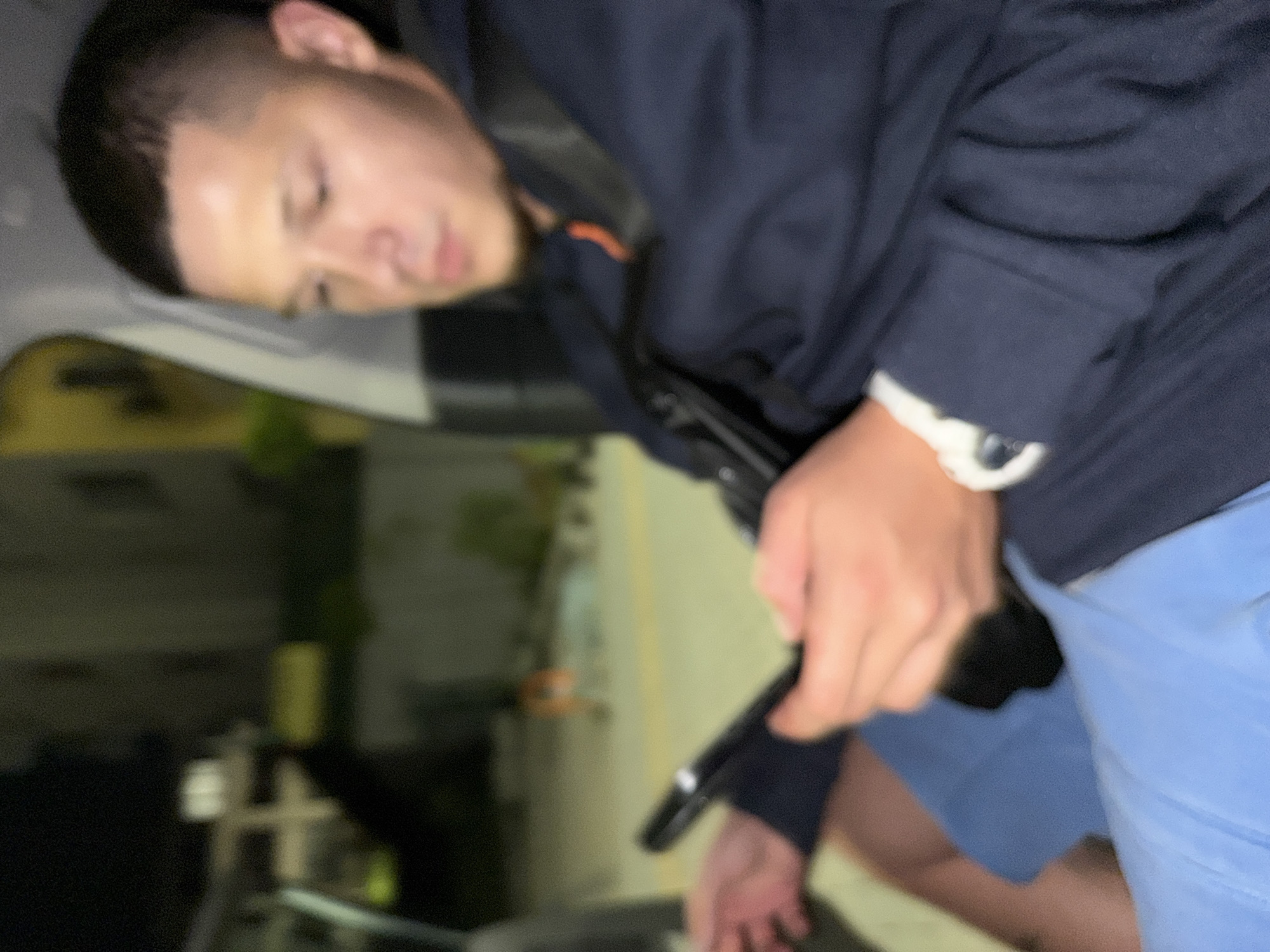 A young man with a fade haircut is sitting in a vehicle, looking at his phone. He is wearing a dark blue jacket with an orange accent at the collar and light blue shorts. His left hand is holding the phone, and his right arm is resting on his lap. The background is blurred but appears to be an outdoor urban setting at night, possibly in Sofia, Bulgaria, with some buildings and streetlights visible. The lighting suggests it is nighttime.