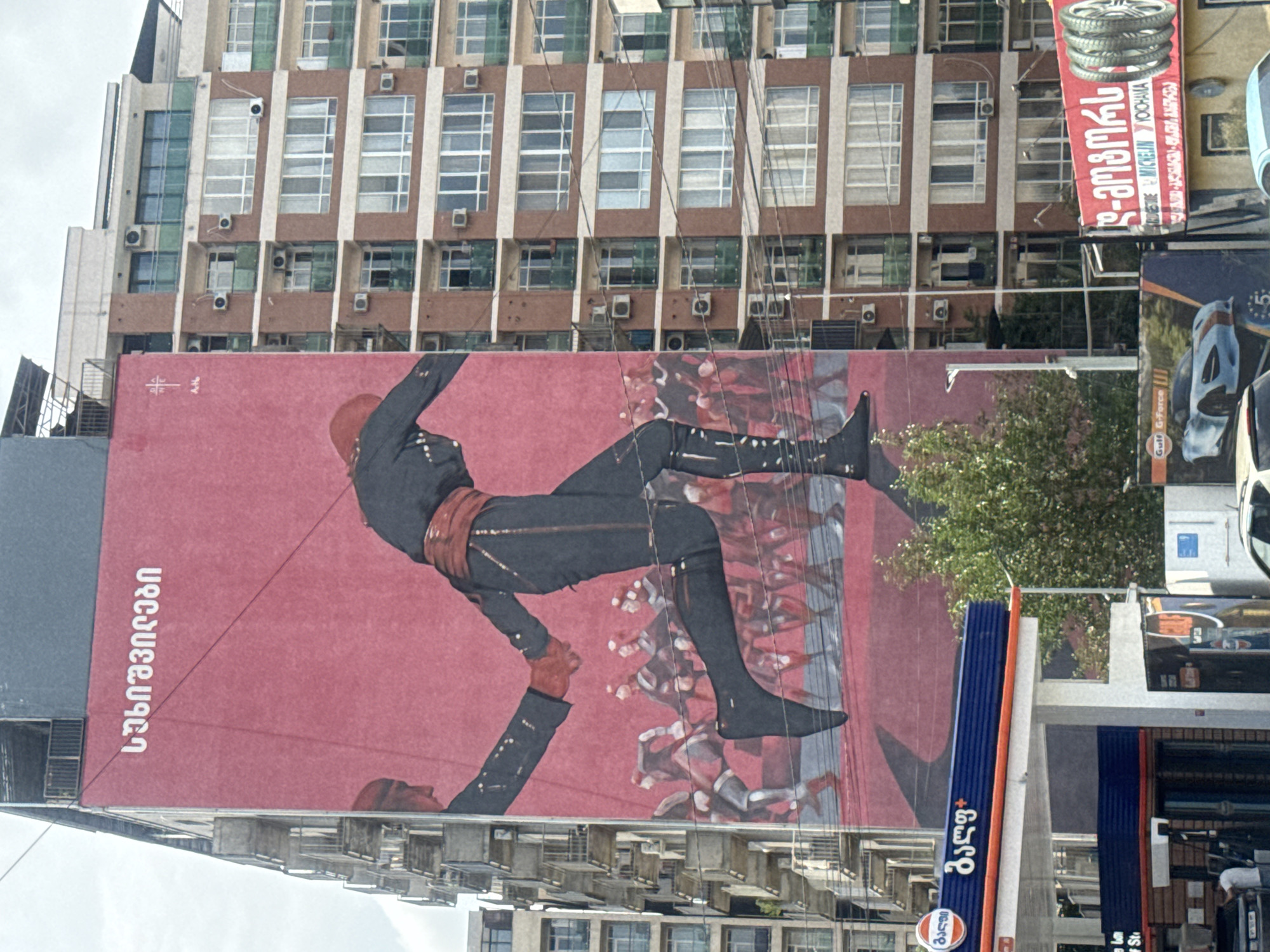 This image captures a mural on the side of a tall, multi-story building in Tbilisi, Georgia. The mural features two figures holding hands, dressed in traditional Georgian attire, with a procession of other similarly dressed figures behind them. The building to the right of the mural is a residential building with numerous windows and balconies. In the foreground, a gas station with a blue and orange facade is visible, along with advertisements for car tires and a sports car. The overall scene depicts a blend of art, urban architecture, and commercial activity.