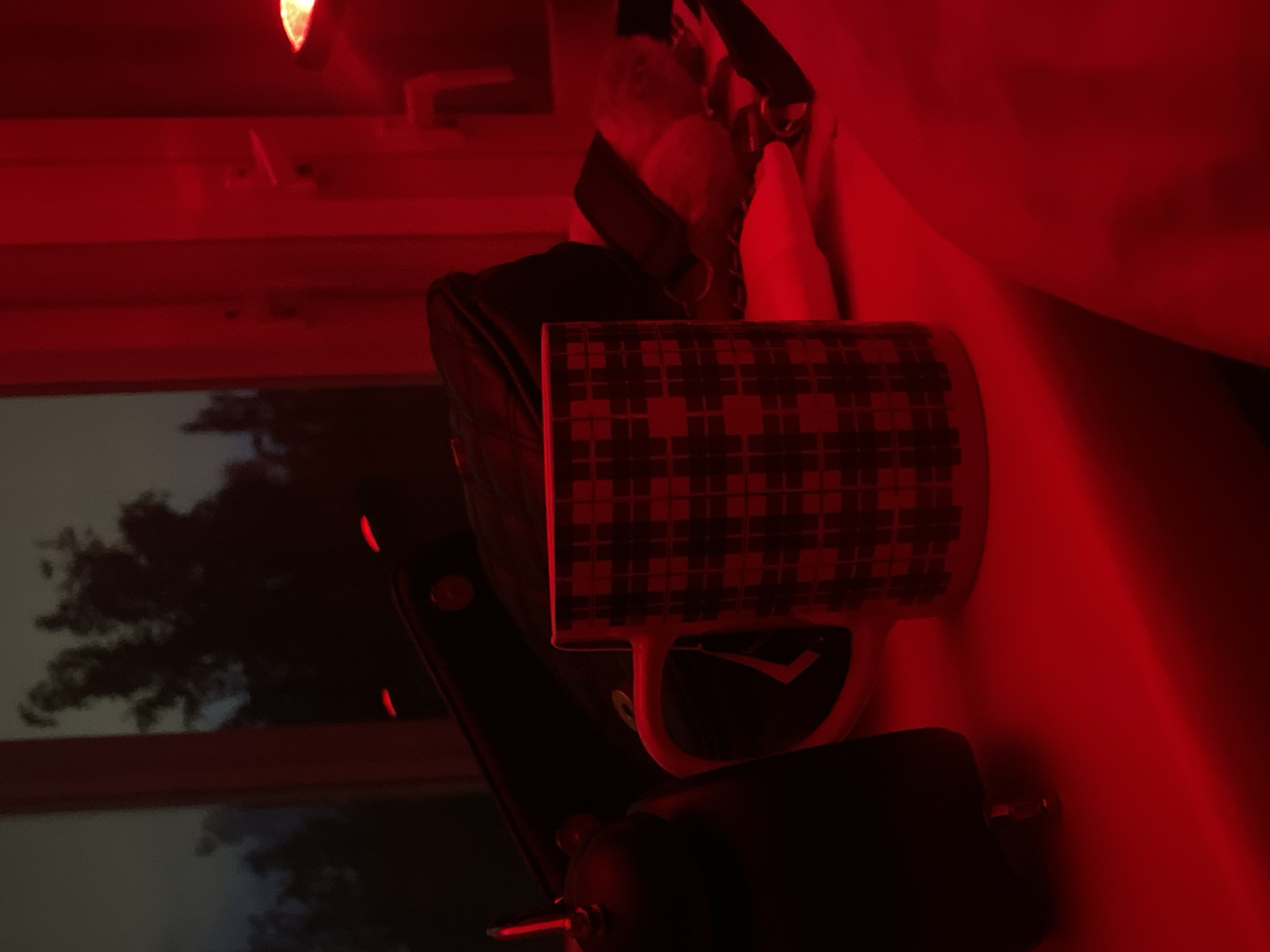 The image is heavily dominated by a strong red light source, casting the entire scene in deep red and dark tones. The perspective is tilted, making objects appear as if seen from above and to the side.
The central subject is a ceramic mug, positioned on what appears to be a flat surface. The mug features a distinct plaid or checkered pattern, composed of darker squares against a background that appears red due to the lighting. A white handle extends from the mug, and a dark "V" shaped logo or symbol is faintly visible on the mug's body near the handle.
To the left of the mug, a dark, soft-sided bag or case, possibly made of leather or fabric, is partially visible. Further back, a window reveals the outdoors, where dark silhouettes of trees are set against a hazy, lighter sky, suggesting it is dusk or night. The window frame itself is light-colored but saturated with red light.
At the top of the frame, a bright, concentrated red light source is visible, illuminating the immediate surroundings. Below the mug, towards the bottom center, a dark, somewhat cylindrical object with a metallic pin or rod protruding from it can be seen.
The scene is an indoor setting, likely a room, characterized by the intense red ambient lighting. No people are present, and no specific activity is taking place; it's a still life. The time of day appears to be evening or night. There is no visible text in the image. The red lighting creates a distinct, moody, and perhaps intimate atmosphere.