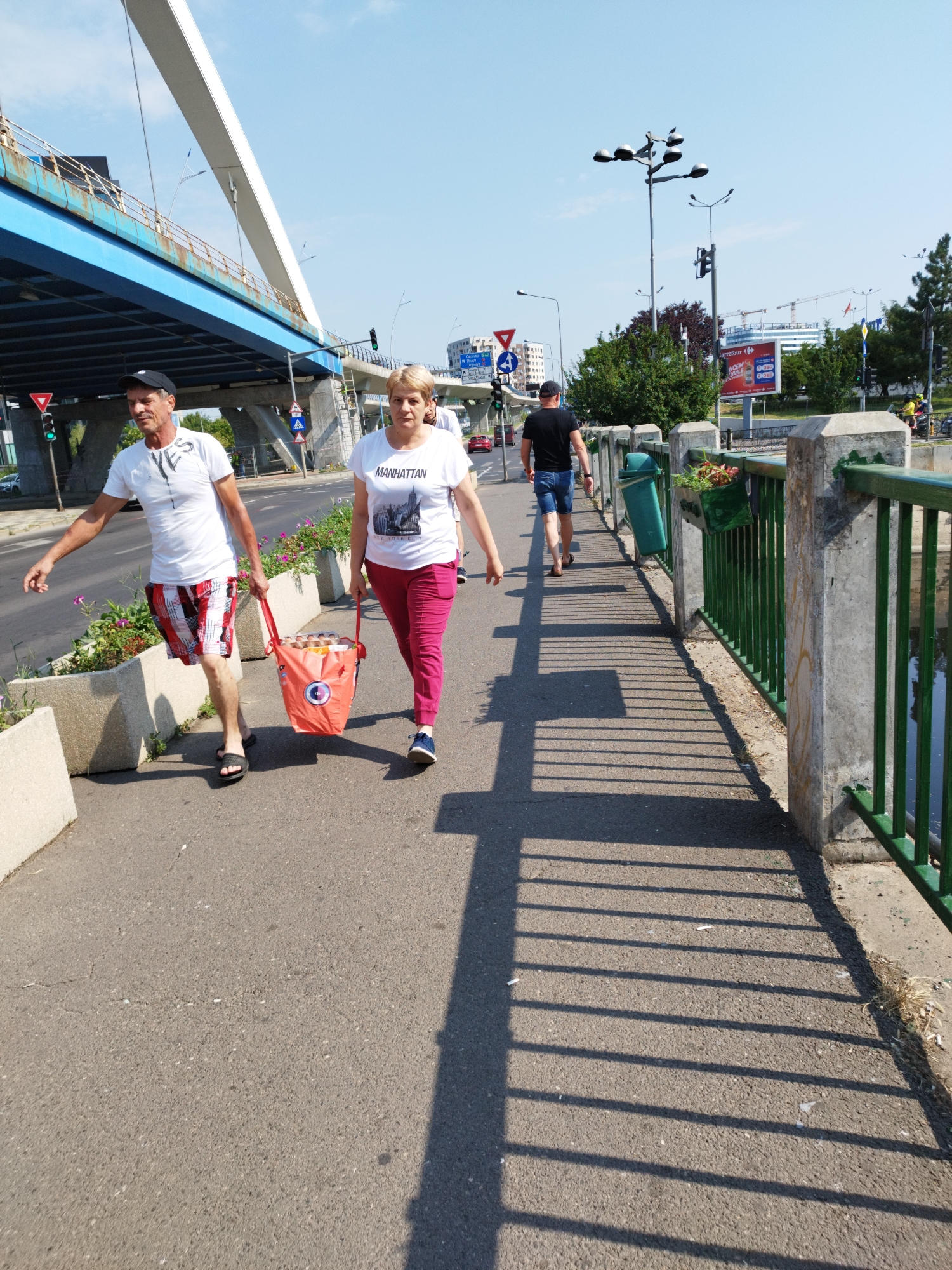 In Bucharest, Romania, a man and a woman walk along a sidewalk next to a bridge overpass. The man, wearing a white t-shirt and red plaid shorts, walks on the left, his right arm extended behind him. He carries a red bag in his left hand. The woman, in a white t-shirt with "MANHATTAN NEW YORK CITY" printed on it and pink pants, walks to his right, also carrying the red bag. The bag appears to contain multiple items, possibly food items. The sidewalk is made of asphalt, and there are planters with flowers along the left side. A green metal railing with concrete posts borders the right side of the sidewalk, overlooking a body of water. In the background, there is traffic, streetlights, and construction cranes, indicating an urban environment. The sky is clear and blue.