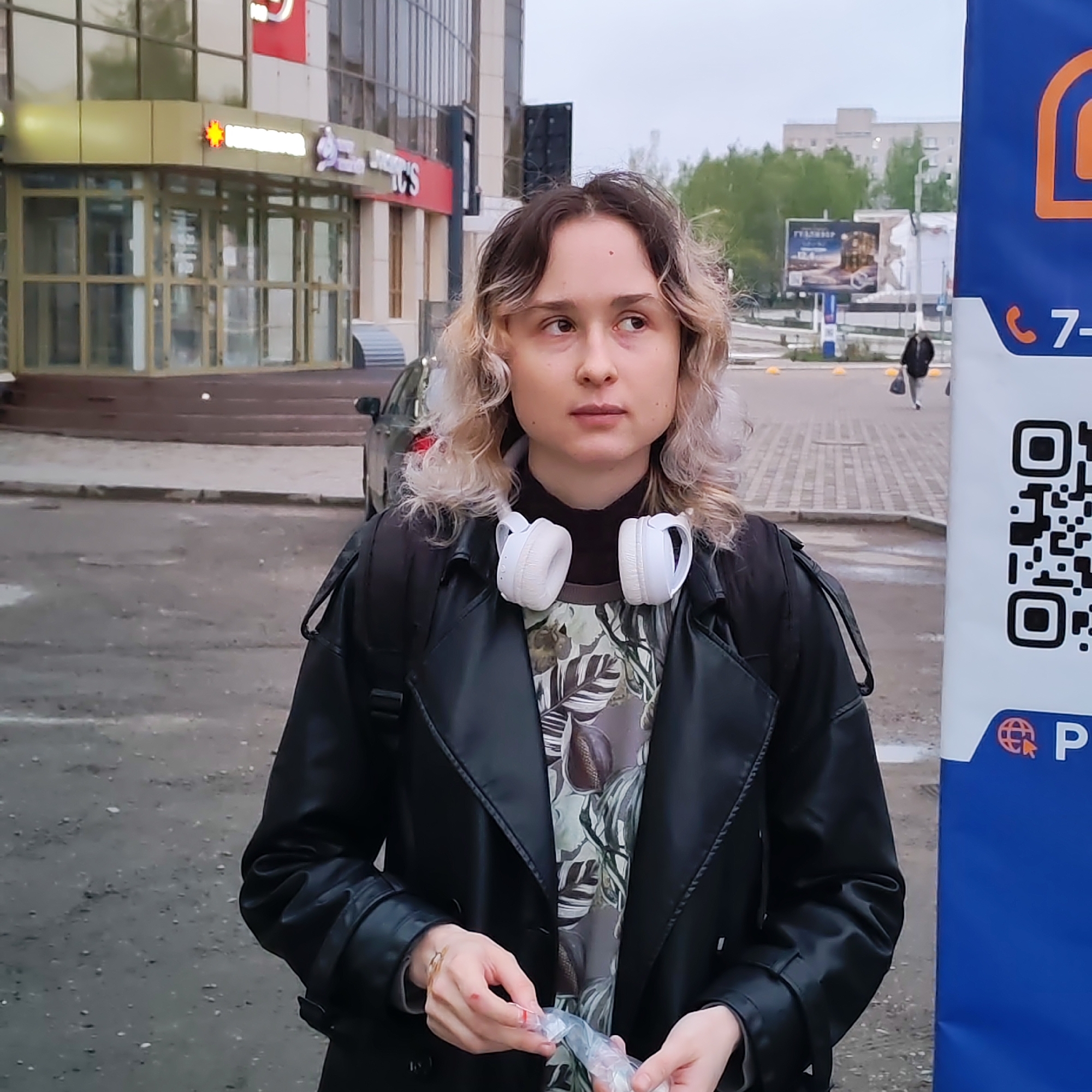 A young woman stands outdoors in Chaikovsky, Russia, near a building with a sign in Russian. She has white headphones around her neck, a black jacket, and is holding a small plastic bag. A blue sign with a QR code is visible to her right. A pedestrian walks in the background toward a building with a billboard.