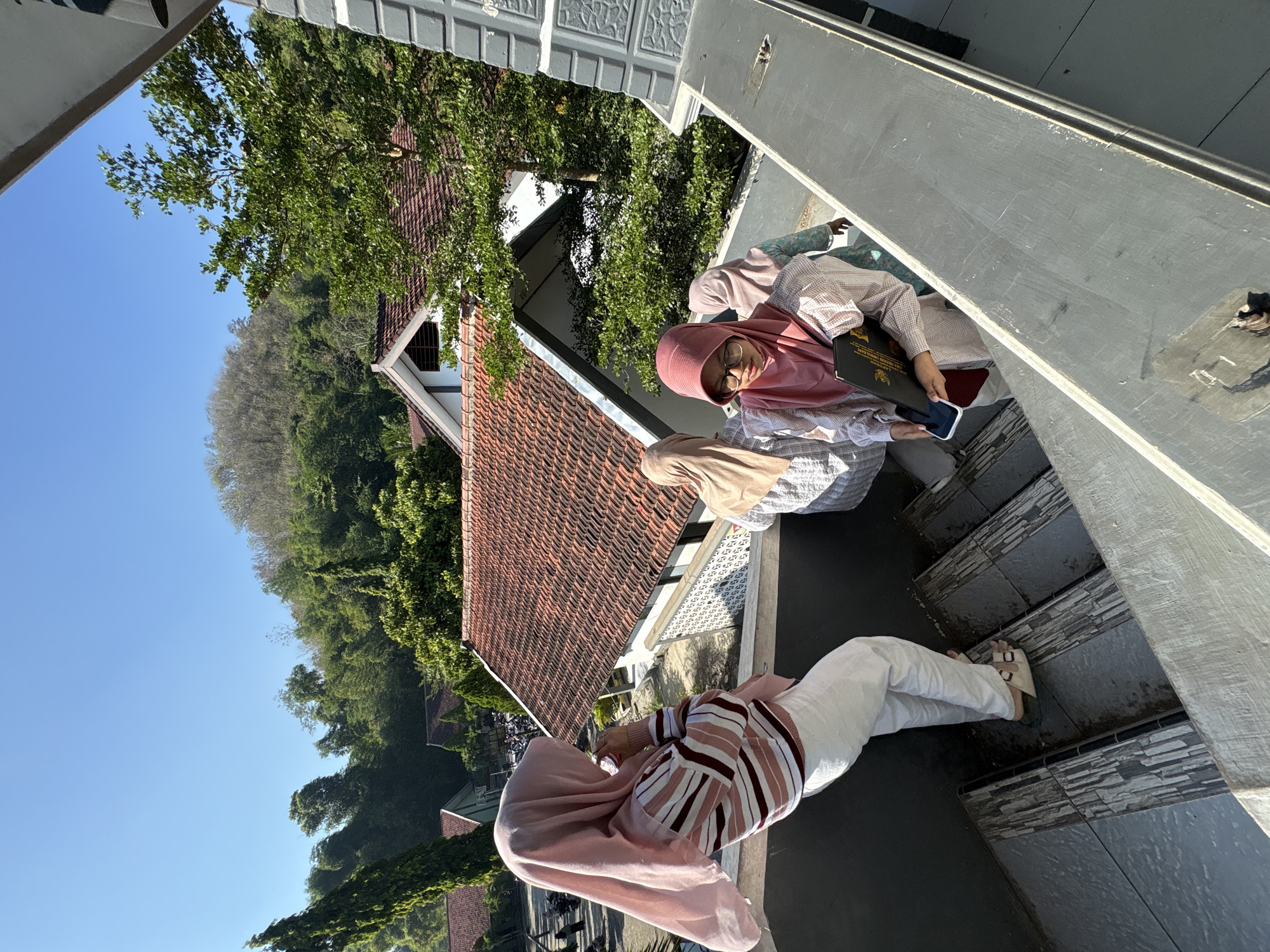 In Gunungsari, Indonesia, on a clear day, four individuals, three of whom are women wearing hijabs, are on an outdoor staircase. The woman in the foreground, wearing glasses and a pink hijab, is looking down and holding a black folder with a government seal. She is wearing a patterned shirt and a maroon undershirt. To her left, another woman in a pink hijab is facing away from the camera, holding a phone to her ear. She is wearing a horizontally striped sweater and white pants. Behind these two women, two other individuals wearing hijabs are visible on the stairs. The staircase leads down from a building with a red-tiled roof, surrounded by lush green trees and a gentle hillside in the background. The stairs themselves have a stone-like pattern.