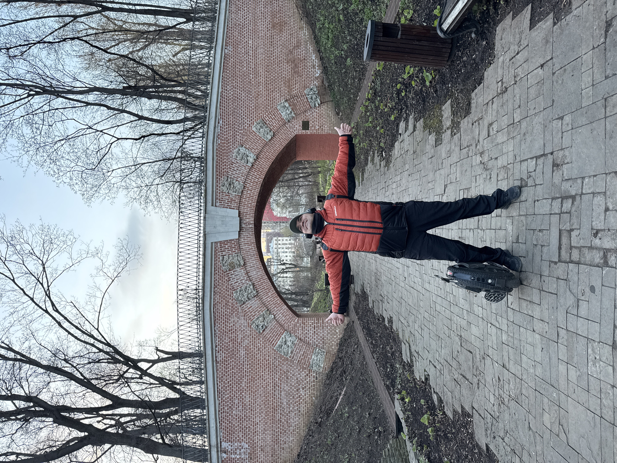 A man in a red and black jacket, black pants, and a dark cap stands on a gray stone-tiled path with his arms outstretched, balanced on an electric unicycle. He is facing the camera. Behind him is a brick archway with a metal railing on top, and through the archway, buildings are visible. Bare trees frame the scene. To the right of the man is a wooden trash bin and a dirt area with sparse vegetation. The location is the Yauza Gates aqueduct, located in Moscow, Russia.