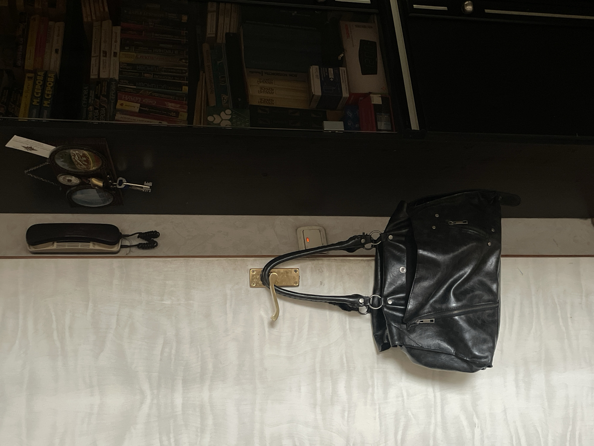 A black leather handbag hangs from a brass door handle on a white paneled door. To the right of the door, mounted on a dark brown cabinet, is an old-fashioned black telephone. Above the telephone, a small shelf displays a framed decorative item containing two miniature oval portraits and a set of keys with a large bell. The cabinet to the right is a tall, dark brown bookcase filled with books and various boxes. The scene appears to be an interior domestic setting, possibly a hallway or entryway, in Moscow, Russia, given the context provided. The lighting is dim, suggesting either late day or a poorly lit interior.