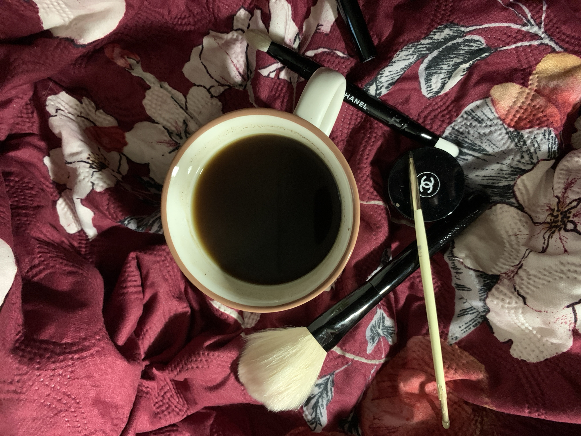 The image depicts a mug of black coffee surrounded by various makeup items, resting on a floral-patterned burgundy fabric. The makeup includes a Chanel eyeshadow pot, a Chanel eyeliner pen, and two makeup brushes: one with a large white brush and another with a smaller brush. Also visible is a wooden applicator tool. The scene is indoors. The location and city are unknown.