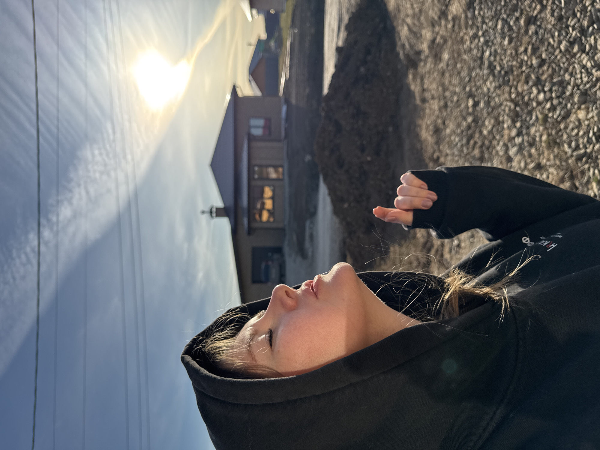 The image, when correctly oriented, captures a young person in the foreground, shown in profile, looking upwards and slightly to their left. They are wearing a black hoodie with the hood pulled up around their face. Their eyes are closed or heavily squinted, possibly due to the intense sunlight. Light brown hair is visible around their face and escaping from the hood. Their right hand is partially visible in the mid-ground, with fingers slightly curled and light pink nail polish on the nails.
The background features a bright, clear blue sky with some scattered wispy clouds. A very strong, almost blinding sun is visible in the upper left of the frame, casting significant glare and light rays. Several utility wires run horizontally across the sky. In the distant background, a building with a light-colored roof and darker walls is visible. The ground surface to the right appears to be a rough, gravel or stony path.
The scene depicts a sunny outdoor setting, likely late morning or afternoon given the strong, direct sunlight. The person appears to be looking towards the sun, perhaps enjoying its warmth or simply reacting to its brightness. On the left shoulder of the black hoodie, partial white text is visible, which includes Cyrillic characters like 'НК' and other indistinct letters. The location context suggests a rural or semi-rural area in Gornyi, Russia.