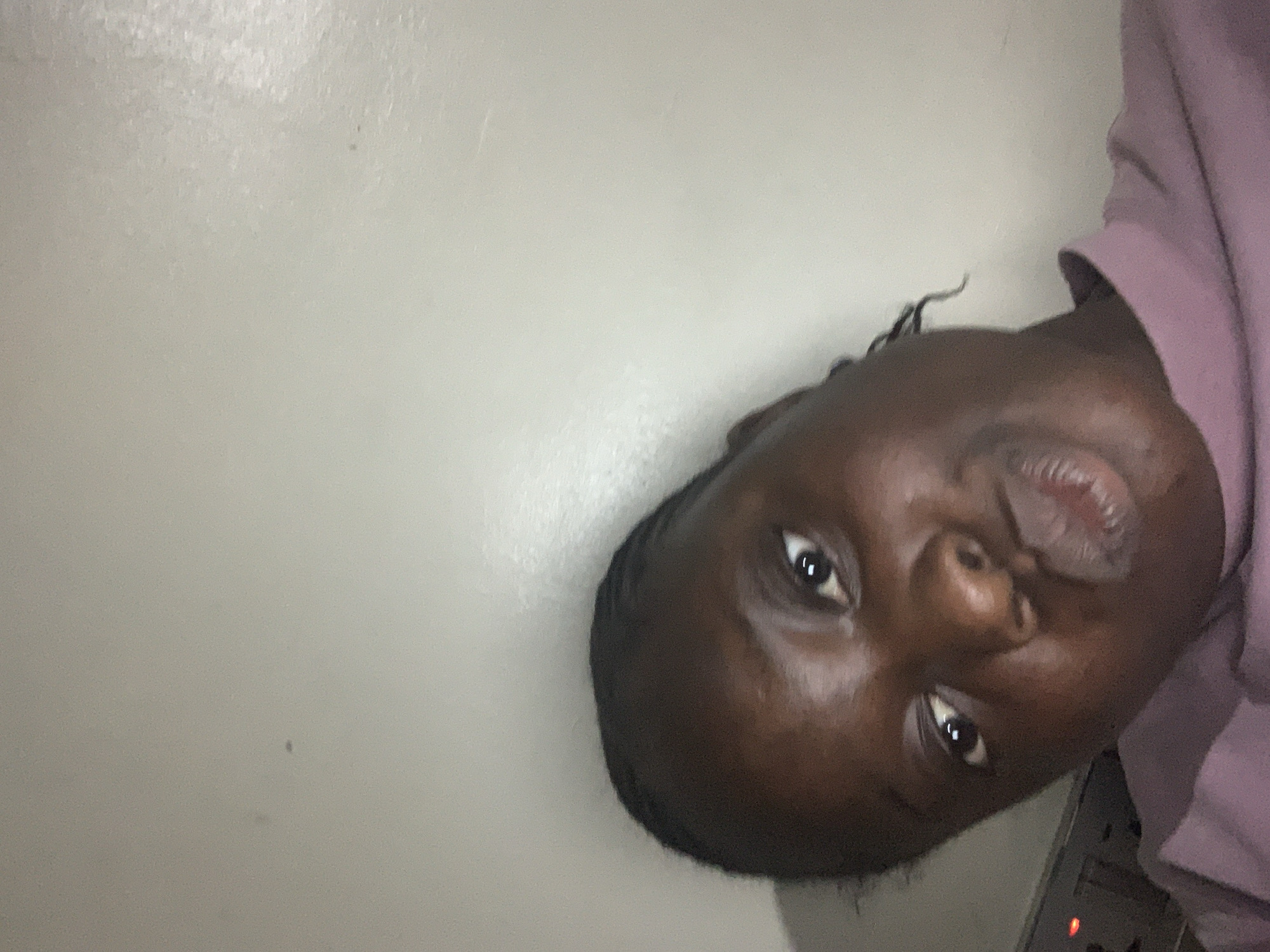 A young Black person is leaning into the camera, making a kissing face. They have braids and are wearing a purple top. The background is a plain, light-colored wall. The image is taken from a low angle.