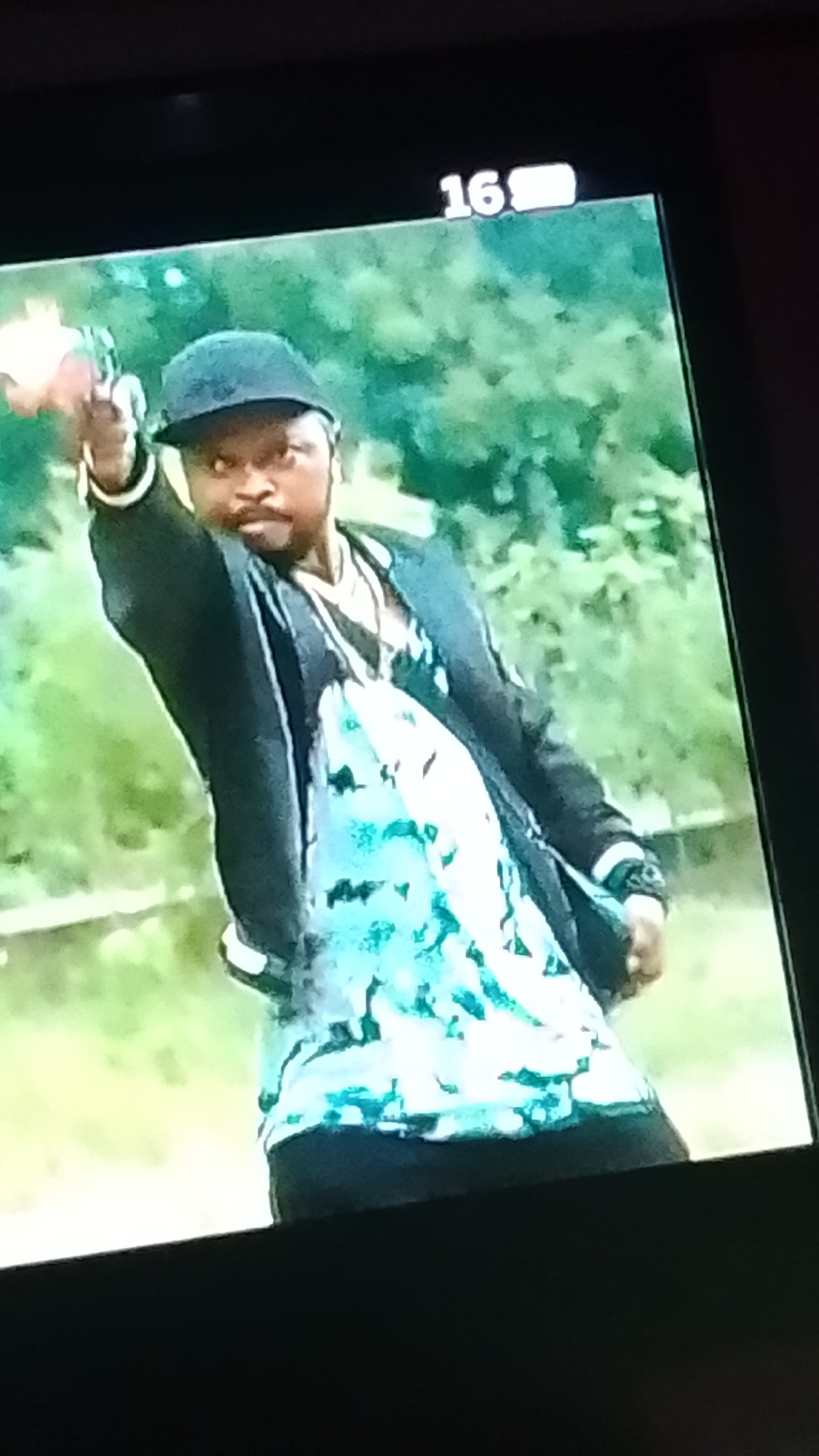 A still image captured from a screen displays a dark-skinned male subject outdoors, purportedly in the city of Lokoja, Nigeria. The subject is positioned facing slightly left of center, with his body angled. He wears a dark baseball cap with the brim forward, a dark open jacket, and a light-colored shirt underneath featuring a prominent teal and white abstract or patterned design. A necklace with a light-colored, possibly silver-toned, pendant is visible. His right arm is extended forward and slightly upward, holding an object resembling a pistol with a light-colored, possibly pinkish, grip. The object is aimed towards the viewer's left. The subject has a beard and a serious or intense facial expression. His left arm is bent, with his hand near his waist. The background consists of blurred green foliage, indicative of a natural outdoor environment. In the upper right corner of the displayed screen, the number "16" is visible in white text, followed by an incomplete battery or signal strength icon.