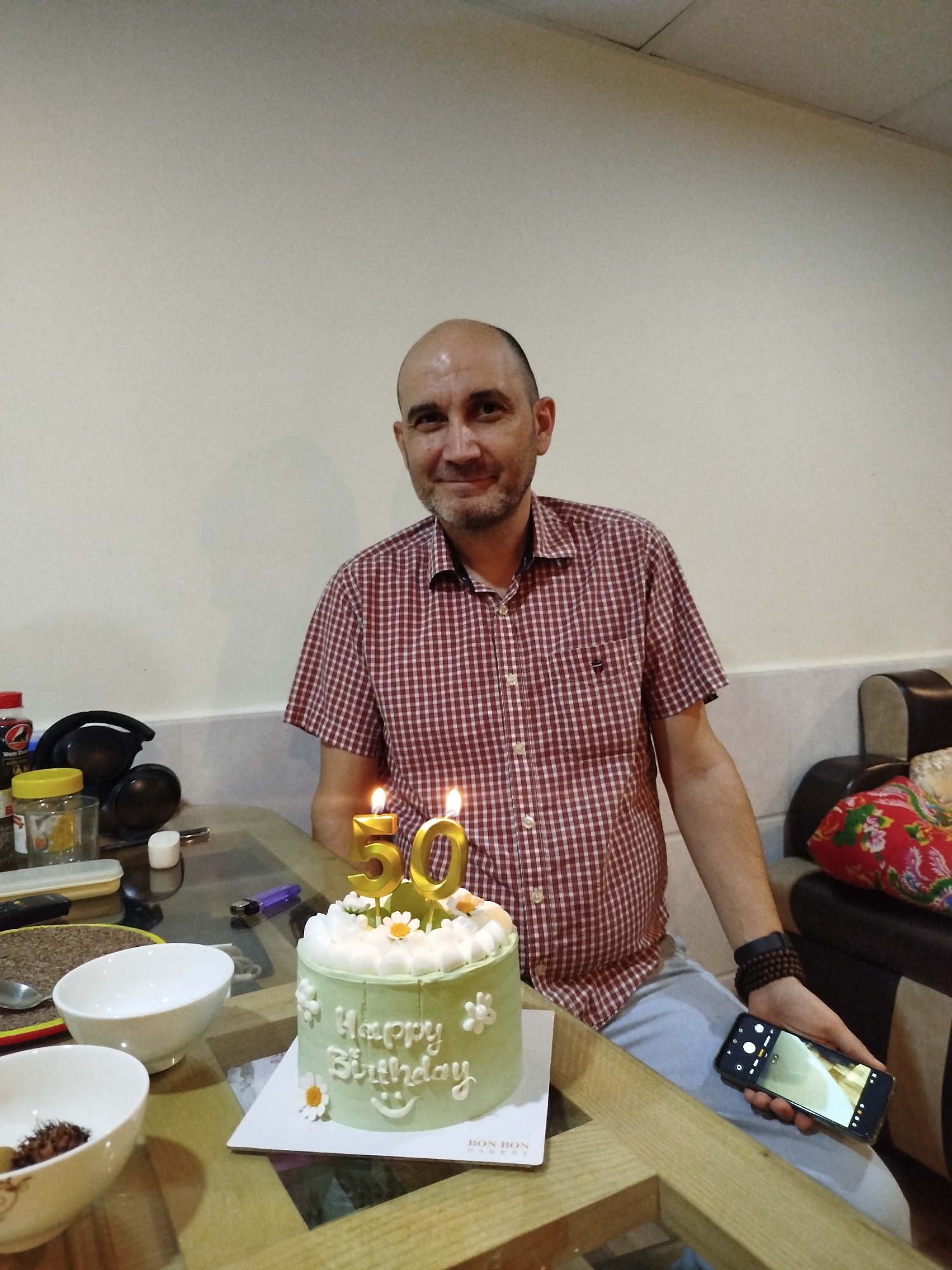 The image depicts a man celebrating his 50th birthday in an indoor setting, likely a home in Da Lat, Vietnam. The central subject is a bald man with a short beard, wearing a red and white checkered short-sleeved shirt and light-colored pants. He is seated at a glass-topped wooden table, smiling and looking directly at the camera.
In front of him is a light green birthday cake, decorated with white frosting swirls and small white flowers. The words "Happy Birthday" are written on the side of the cake in white frosting. Two golden candles, shaped as the numbers "50," are lit and placed on top of the cake. The cake sits on a white board with "BON BON" printed on it.
On the table, several items are visible: two white ceramic bowls (one empty, one containing a snack), a black pair of headphones, a yellow-lidded jar, a red bottle, and a purple lighter. The man holds a smartphone in his left hand, its screen showing a live camera view, suggesting he might be recording or taking another photo. He also wears a dark, multi-strand bracelet on his left wrist.
The background consists of a plain light-colored wall with a white baseboard, and a partial view of a dark sofa with a red and white floral patterned cushion. The lighting suggests an indoor setting, possibly evening or night, using artificial light. The man's smiling expression indicates happiness and enjoyment of the event.