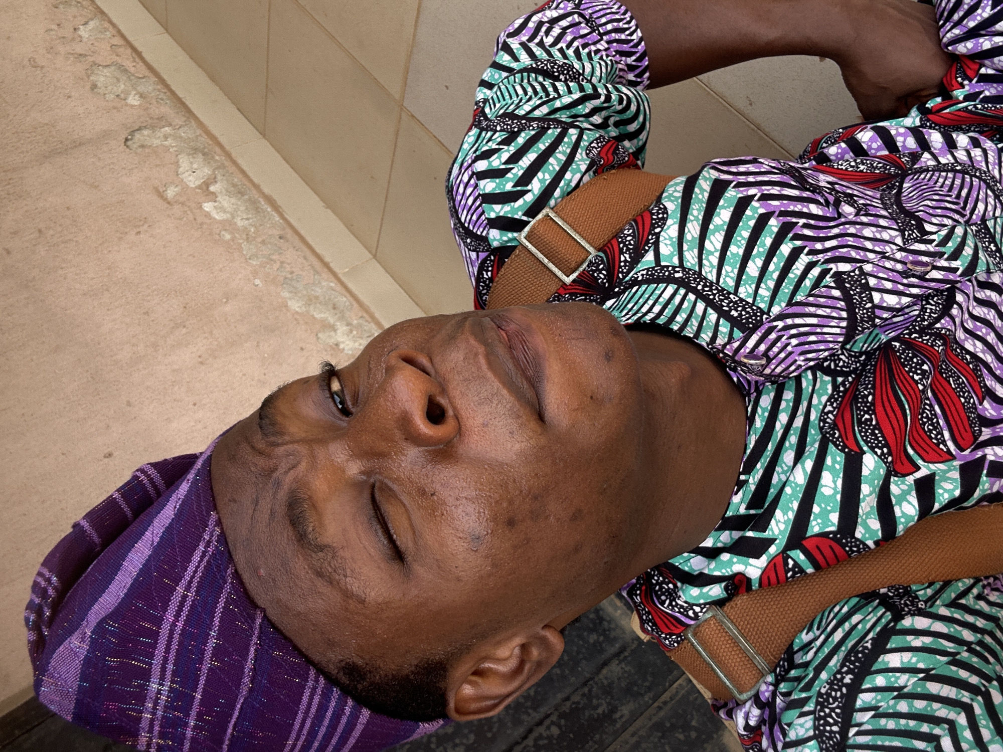 A medium shot shows a dark-skinned person in the city of Unknown, Unknown, possibly resting. They are wearing a patterned shirt with purple, green, red, and black designs, along with a purple striped head covering. A brown strap with a silver buckle crosses their chest. The person's eyes are mostly closed, and their head is tilted back. The background includes a neutral-toned surface, possibly concrete or tile.