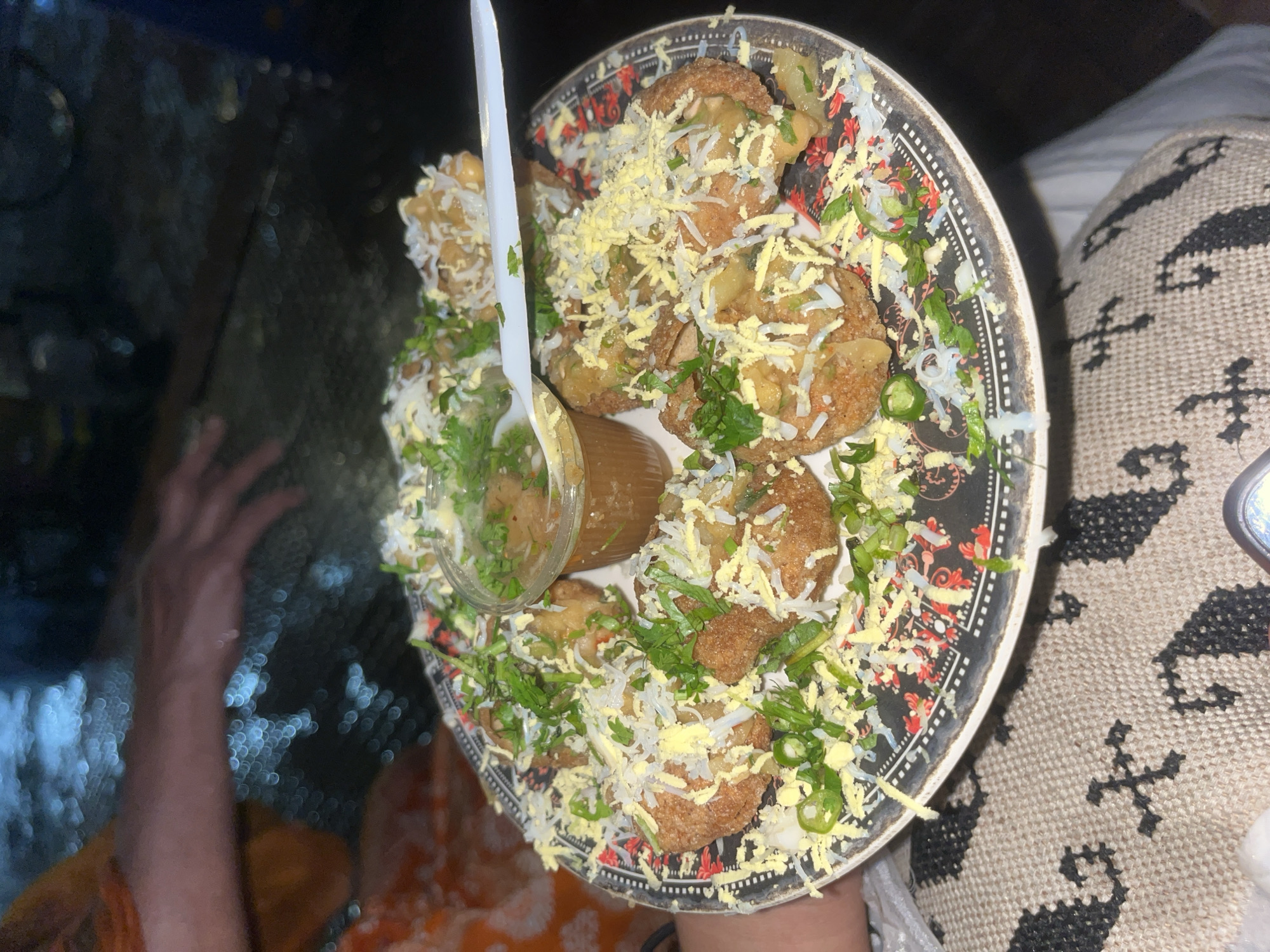 A top-down, close-up shot shows a plate of pani puri, a popular street food, held in someone's hands. The pani puri is topped with shredded cheese, chopped green chilies, cilantro, and white noodles. A small plastic cup filled with tamarind water and a white plastic spoon sits on the side of the plate. The plate has a black background with a decorative border. The person holding the plate is wearing a sweater with a black and white pattern. The image has a slightly dark and grainy quality. Based on the food and context, it is likely taken in Dhaka, Bangladesh, where pani puri is a common street food item.