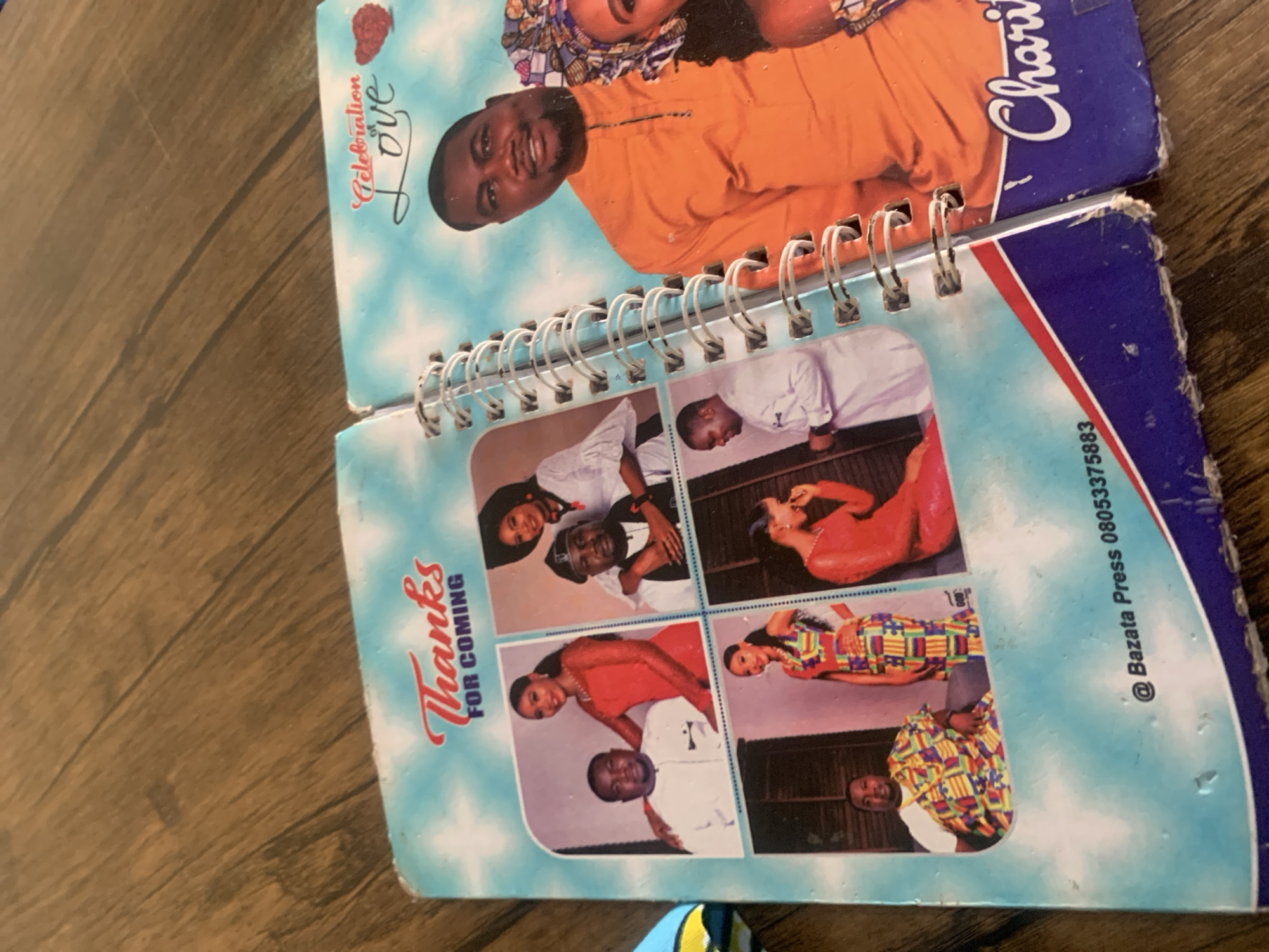 The image shows an open spiral-bound booklet or journal. The left page has the words "Thanks FOR COMING" printed in red script. Below this are four smaller framed photographs: a couple in traditional attire, a man and woman in white outfits, a woman in a colorful dress, and a woman in a red dress. The right page has the words "Celebration Love" in red script with a red heart graphic. A larger photograph on this page shows a man in an orange traditional outfit and a woman in a patterned headwrap. The bottom of the right page has the word "Charity" in white script. The text "@ Bazata Press 08053375883" is visible on the bottom of the left page. The booklet is resting on a wooden surface, likely a table, with a visible wood grain pattern. The setting appears to be indoors. Based on the contact information, the booklet was likely produced by Bazata Press in Nigeria. The text "Jauro Yinu, Nigeria" is not present in the image.