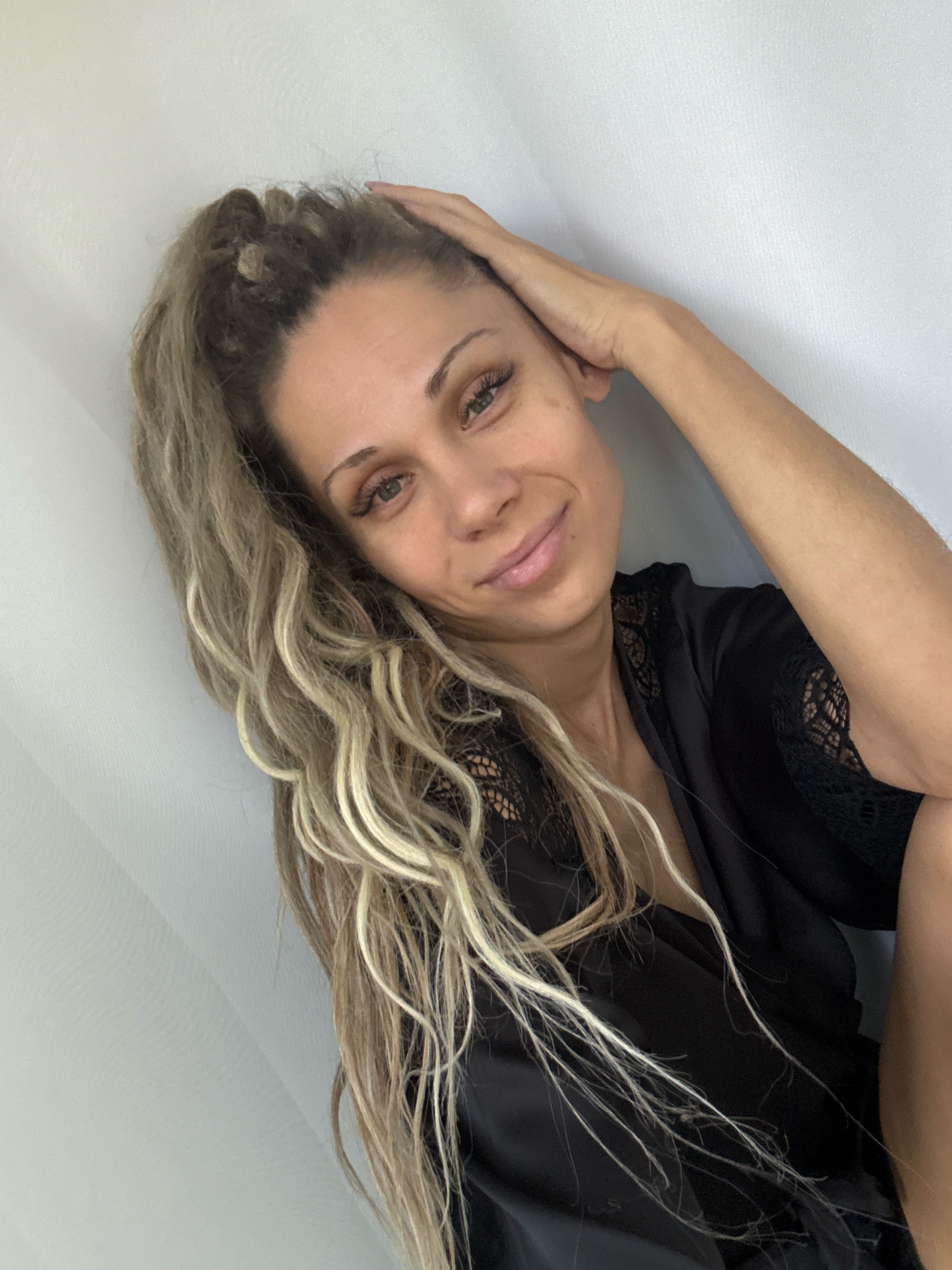 A woman with blonde, wavy hair pulled into a high ponytail, wearing a black robe with lace trim, poses with her hand on her head. She is looking at the camera and smiling. The background is a plain, light-colored wall. The location is not specified in the provided context.