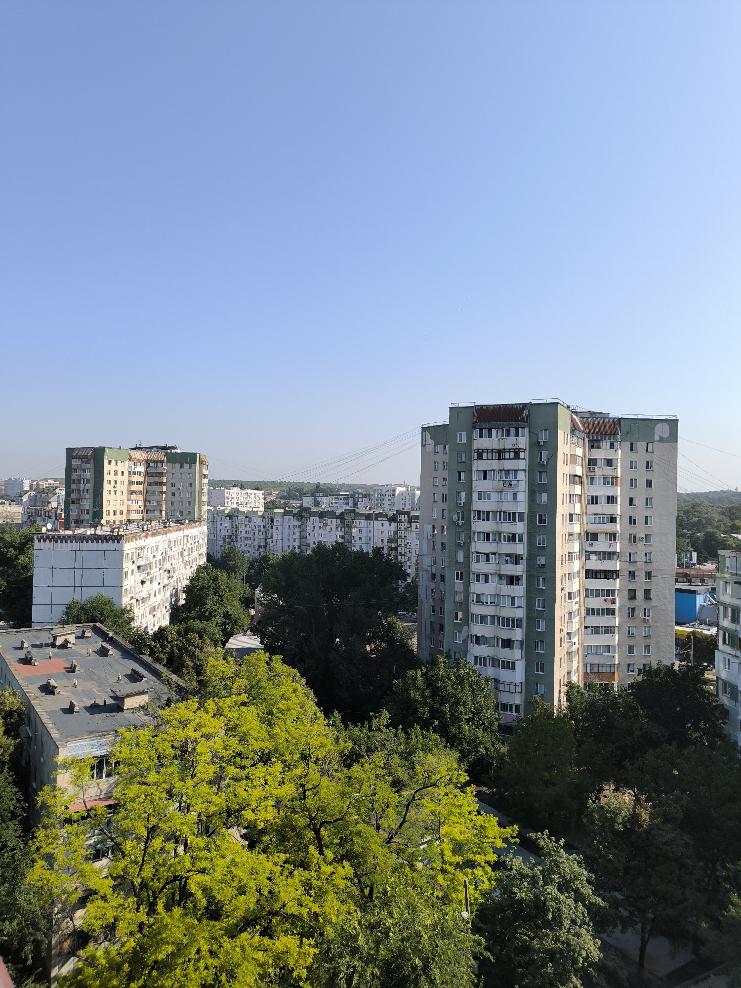 This is a high-angle, wide shot of a residential neighborhood in Chișinău, Moldova, on a clear, sunny day. The scene is dominated by Soviet-era apartment buildings and lush greenery.
In the foreground, a multi-story apartment building with a flat, dark roof is visible, featuring several vents and what appear to be skylights or access hatches. To the left and slightly behind it, another apartment building with a light-colored facade and a row of balconies is partially obscured by trees.
The mid-ground features two prominent apartment buildings. The one on the left is a long, light-colored building with a green-trimmed top and numerous balconies. The one on the right is taller and more imposing, with a predominantly light-colored facade and green accents near the roof. This building has many windows and balconies, and air conditioning units are visible on some of them.
In the background, a cluster of similar apartment buildings stretches towards the horizon. The sky is a clear, pale blue. Dense green trees fill the spaces between the buildings, with some trees in the foreground exhibiting bright, yellow-green foliage, suggesting it might be late spring or early summer. Power lines crisscross the sky in the distance. The overall impression is of a densely populated urban area with a significant amount of green space integrated within it. There are no people or vehicles visible in the frame. The light suggests it is daytime, likely late morning or early afternoon, given the sharp shadows cast by the buildings.