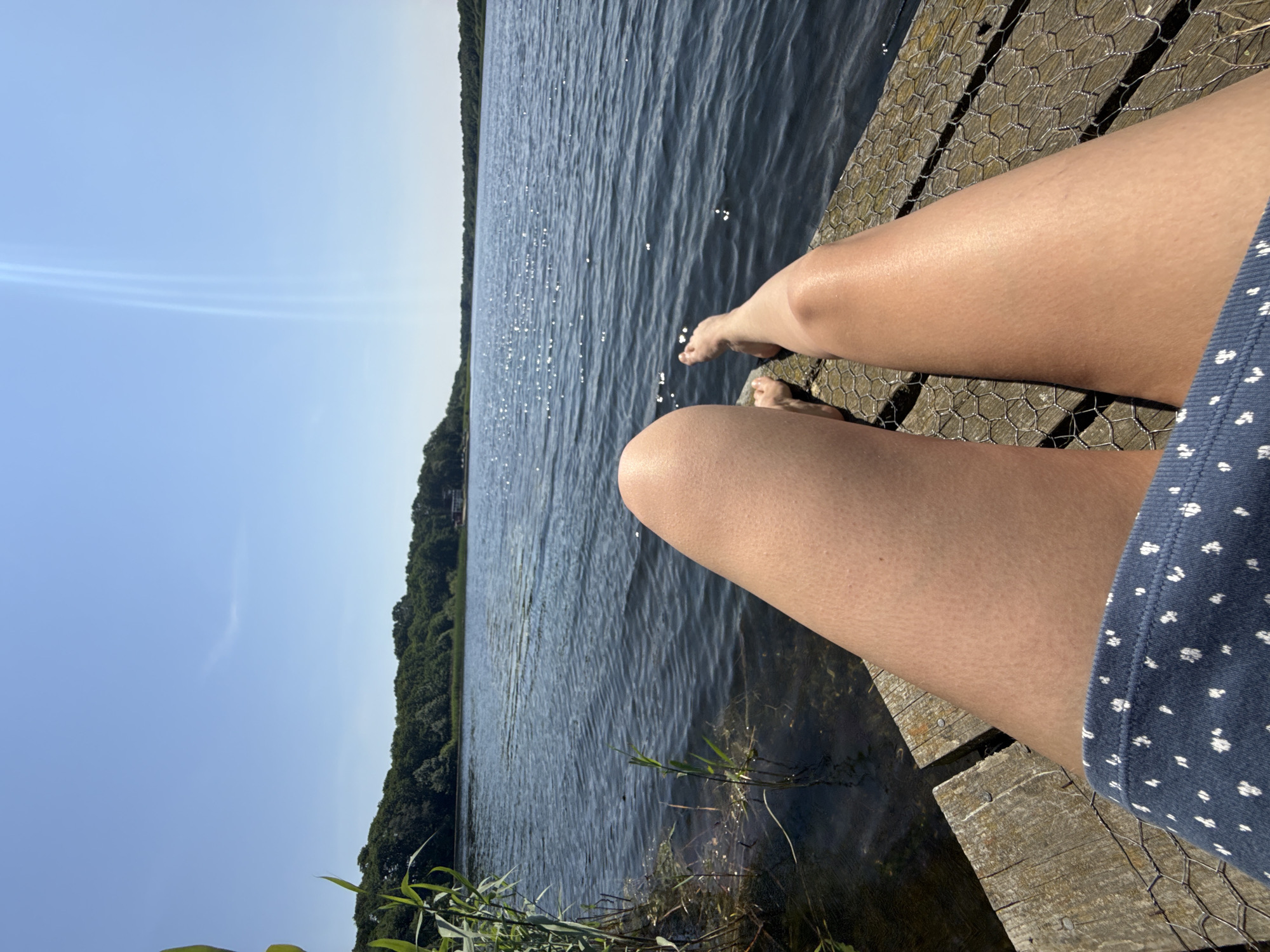 The image shows a close-up view of a person's legs and feet resting on a wooden dock. The person is wearing blue shorts with a white polka dot pattern. The dock is weathered and has a wire mesh covering. The background features a calm lake or body of water, with the surface reflecting sunlight. Beyond the water, a lush green treeline forms the horizon, under a clear blue sky. There are reeds growing along the edge of the water near the dock. The scene appears to be outdoors on a sunny day. The location is not identifiable as being in Great Yarmouth, United Kingdom, or any specific city.