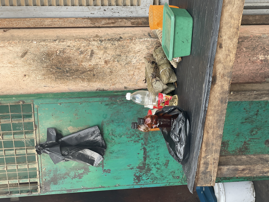 The image displays an assortment of items arranged on and around a weathered wooden surface, suggesting an outdoor or informal setting in Luanda, Angola.
The primary collection of objects rests on a rough, dark wooden plank. On one end, there are two plastic containers: one orange (partially visible) and one green, both rectangular with lids. Beside these are several bundles wrapped in what appear to be plant leaves, tied with string, resembling traditional food preparations like tamales. Further along the plank, a clear plastic bottle with a red and yellow label is visible, next to a brown plastic bottle that is partially covered by a black plastic bag. Another crumpled black plastic bag, containing more brown bottles, lies nearby.
The wooden plank is positioned over a green, metallic surface that shows significant signs of rust and wear, possibly a large generator, utility box, or a sturdy metal table. This green surface also features a rectangular metal grate on one side. A crumpled black plastic bag is also present on this green surface. The background of the scene is a rough, light brown or grey concrete wall. Below the wooden plank, part of a white bucket with a blue handle can be seen.
The arrangement of objects suggests they could be goods laid out for sale at an informal market stall or simply items in temporary storage. No people are visible, and no specific activity is actively taking place. The overall impression is one of a functional, somewhat rustic outdoor space. The items and surfaces are worn, reflecting exposure to the elements. No clear, legible text is visible on any of the items or surfaces. The lighting indicates it is daytime.