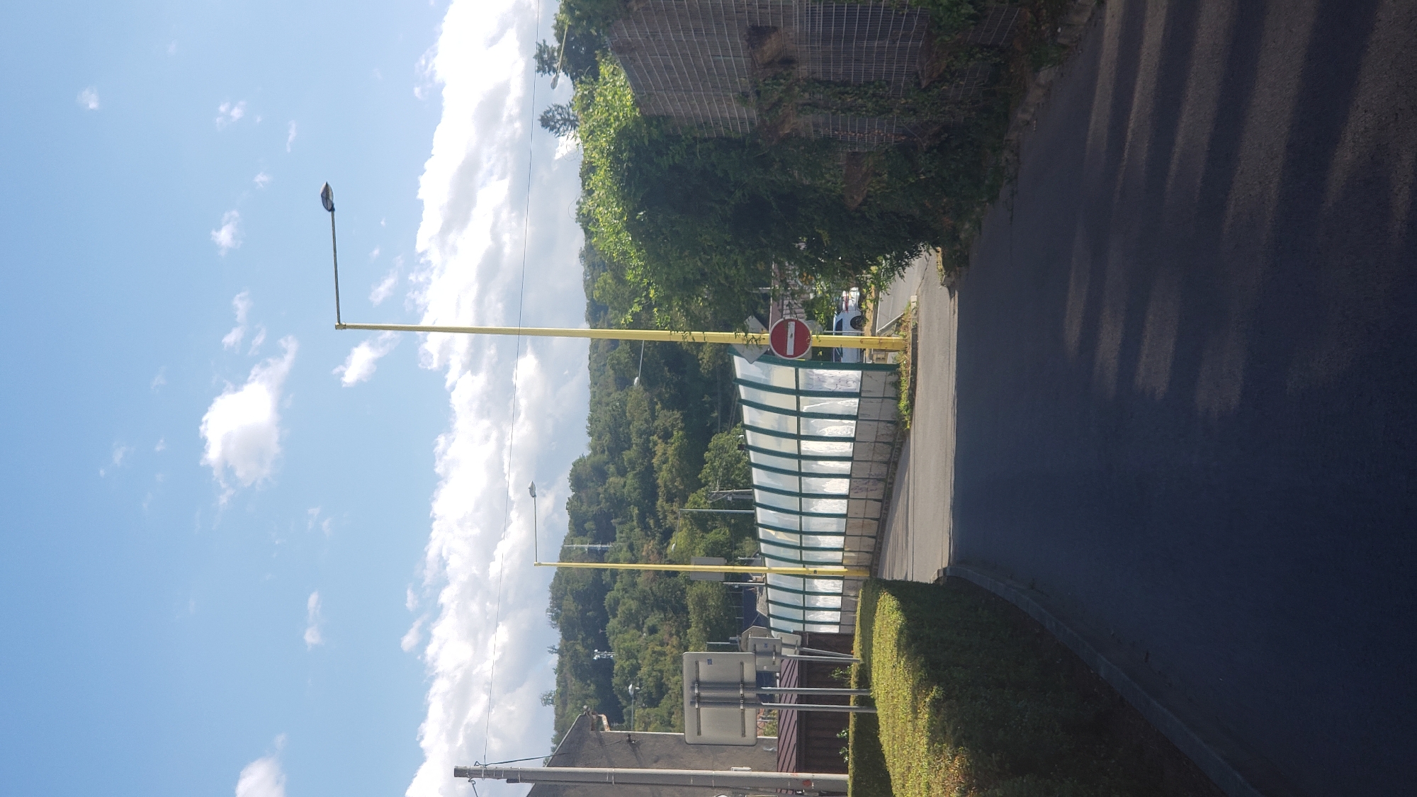 The image captures a view of a road leading towards a covered walkway or tunnel structure, likely a noise barrier, on a sunny day in Prešov, Slovakia. The walkway features a transparent, curved roof supported by green frames and appears to have graffiti on its lower section. To the left of the road, a neatly trimmed green hedge lines the walkway. Two tall yellow lampposts stand near the walkway; one is positioned on the left and the other on the right. A red and white circular "no entry" sign is visible on the right lamppost. In the background, a wooded hillside rises, dotted with trees and some distant buildings. The sky is blue with scattered white clouds. The road surface is asphalt. A white car is partially visible behind the noise barrier. The image appears to have been taken from a low vantage point, looking up slightly towards the covered structure and the hillside.