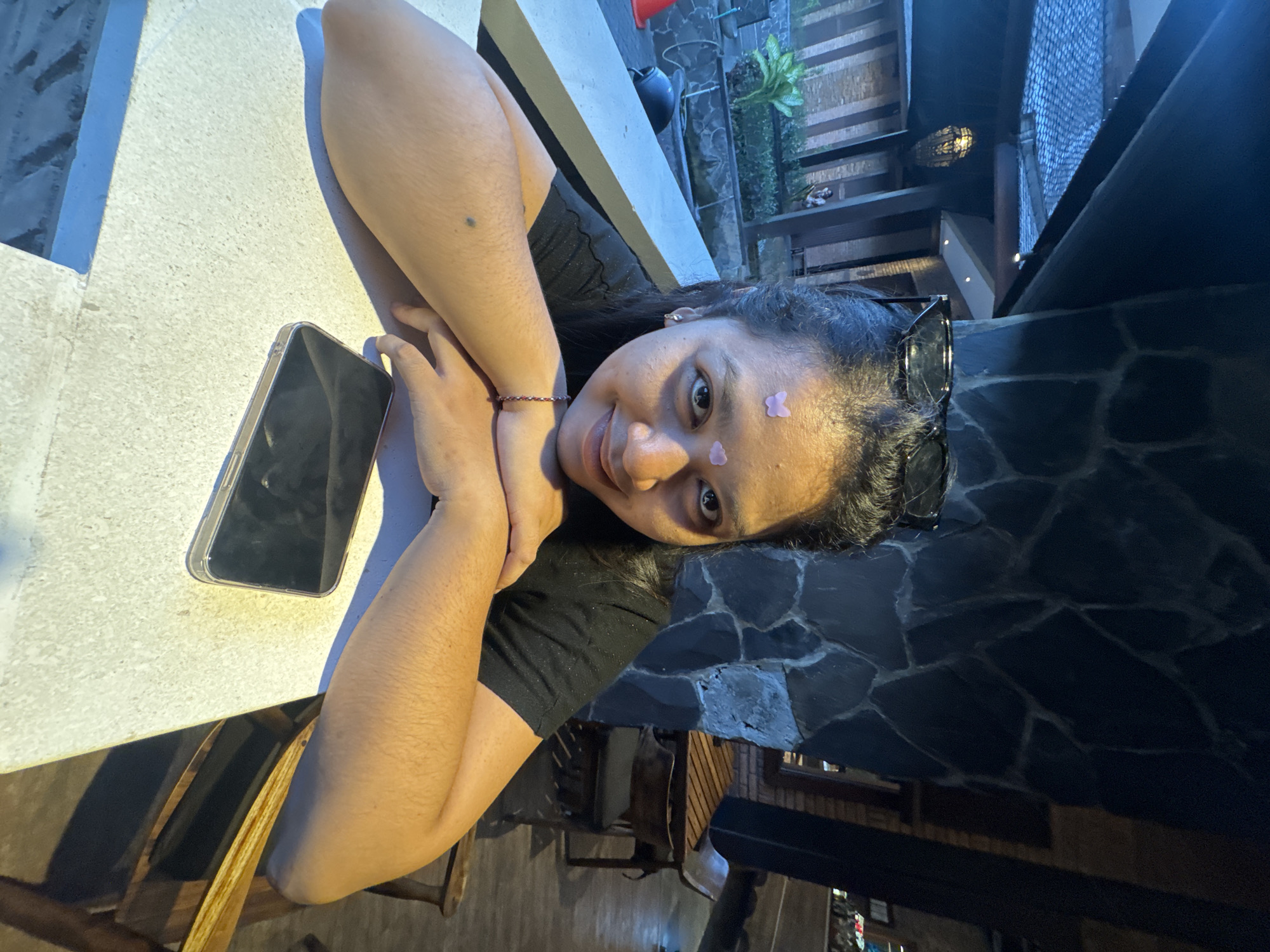 A young woman poses for a photo in what appears to be an outdoor seating area of a restaurant or hotel, possibly in Kuta Selatan, Indonesia. She is resting her head on a black stone table, with her arms folded. A smartphone is placed on a white table nearby. She has a small butterfly sticker on her forehead. Sunglasses are pushed up on her head. The background includes a wooden structure and some plants. The lighting is somewhat dim, suggesting it might be evening.