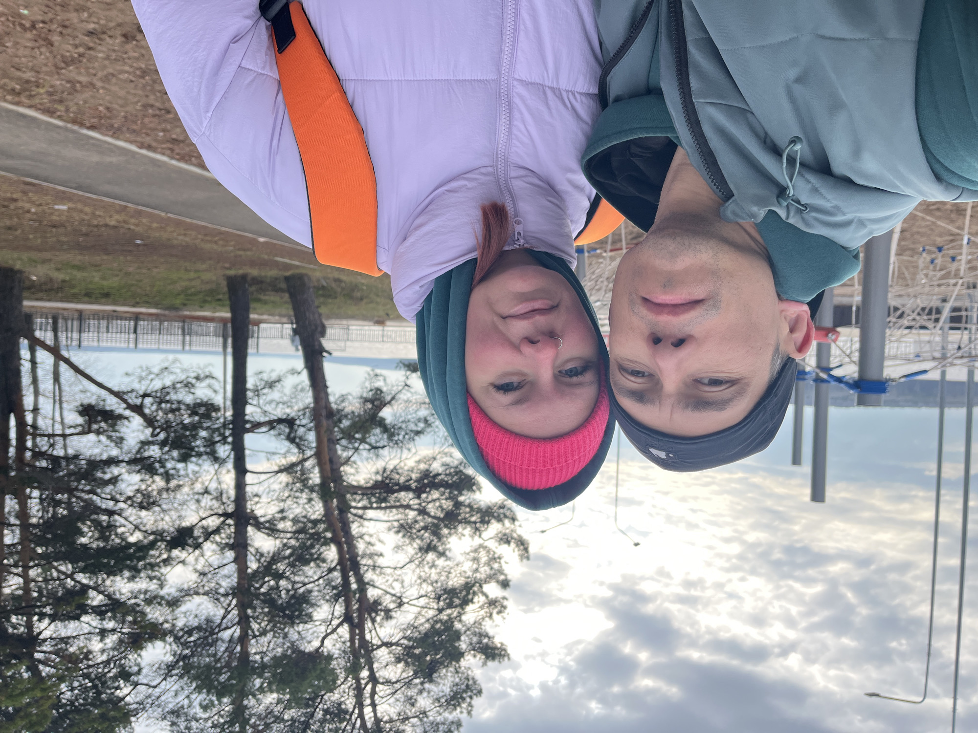 Two people, a woman and a man, are posing for a selfie outdoors. The woman is wearing a pink knit hat under a green hooded jacket, and a light purple puffer jacket with orange straps. The man is wearing a dark beanie and a green jacket.
The background features trees, a body of water, and cloudy skies. The location is identified as the city of Chaikovsky, Russia, although specific landmarks or street names are not discernible from the image.