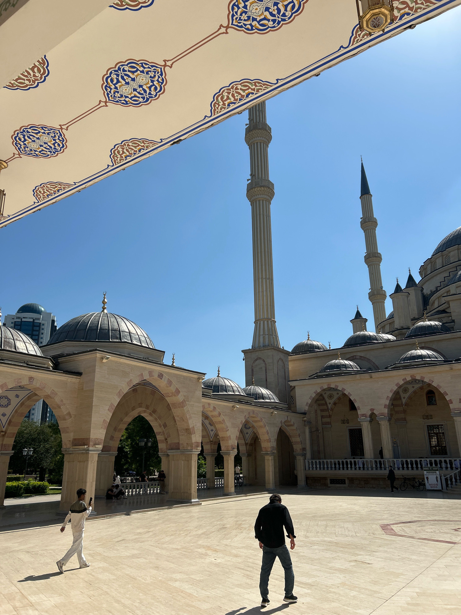 This is a medium shot, eye-level, outdoor, daytime photograph of the Akhmad Kadyrov Mosque in Grozny, Russia. The mosque features a large courtyard paved with light-colored stone, with a colonnade of arched walkways running along one side of the courtyard. The mosque's domes and minarets are visible in the background, against a clear blue sky.
In the foreground, two men are walking in the courtyard. One man, on the left, is wearing a white tracksuit and appears to be looking at his phone as he walks. The other man, on the right, is wearing a black shirt and jeans, and is walking away from the camera.
The architecture of the mosque is characterized by its ornate decorations, including intricate geometric patterns on the ceiling of the covered walkway in the foreground, and the crescent moon finials atop the domes and minarets. The overall impression is one of grandeur and religious significance.