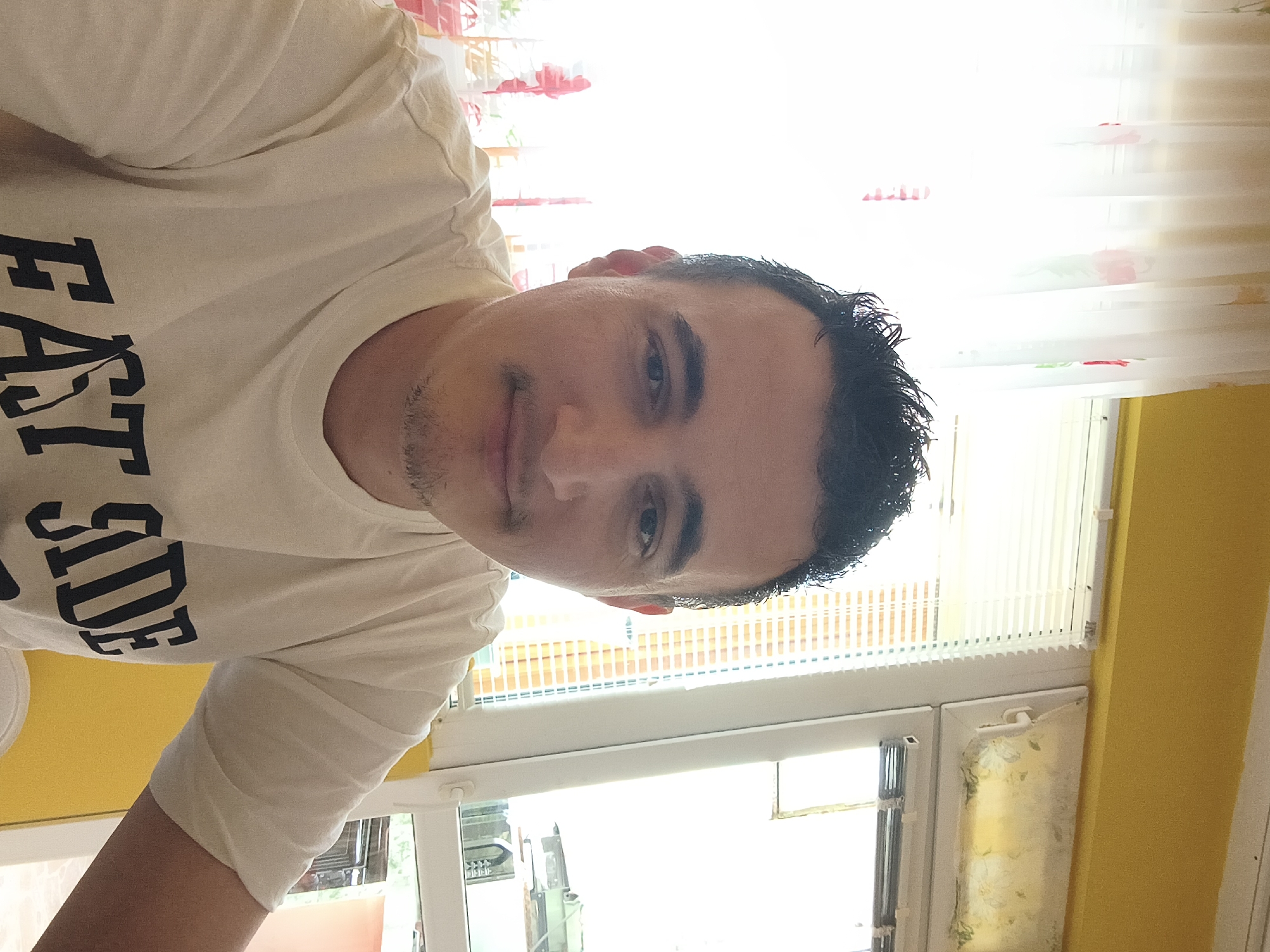 The image depicts a male individual positioned indoors, identified as being located in Shumen, Bulgaria.
The subject is a young adult male, likely in his late 20s to early 30s, with fair skin. He has short, dark, textured hair styled with some volume on top. His facial features include dark eyes, thin eyebrows, and a thin mustache connected to a short chin strap beard. He is looking directly at the camera with a subtle, closed-lip smile. He is wearing a cream or off-white short-sleeved t-shirt with the dark, block letters "EAST SIDE" prominently displayed across the chest.
The background consists primarily of a large window area. To the subject's left, a sheer white curtain with vertical floral patterns in shades of red/pink and green is visible, filtering bright natural light. To the subject's right, a white UPVC window frame is prominent. The left pane of this window features vertical blinds, while the right pane has a frosted or patterned film with faint green and yellow details. A white window handle is visible on the right pane. Above the window frames, a brightly painted mustard yellow wall is present. Through the lower right section of the window, blurred outdoor elements are discernible, including green foliage, possibly a tiled roof, and a peachy-colored exterior wall of another structure. The scene is brightly lit by natural light emanating from behind the subject.