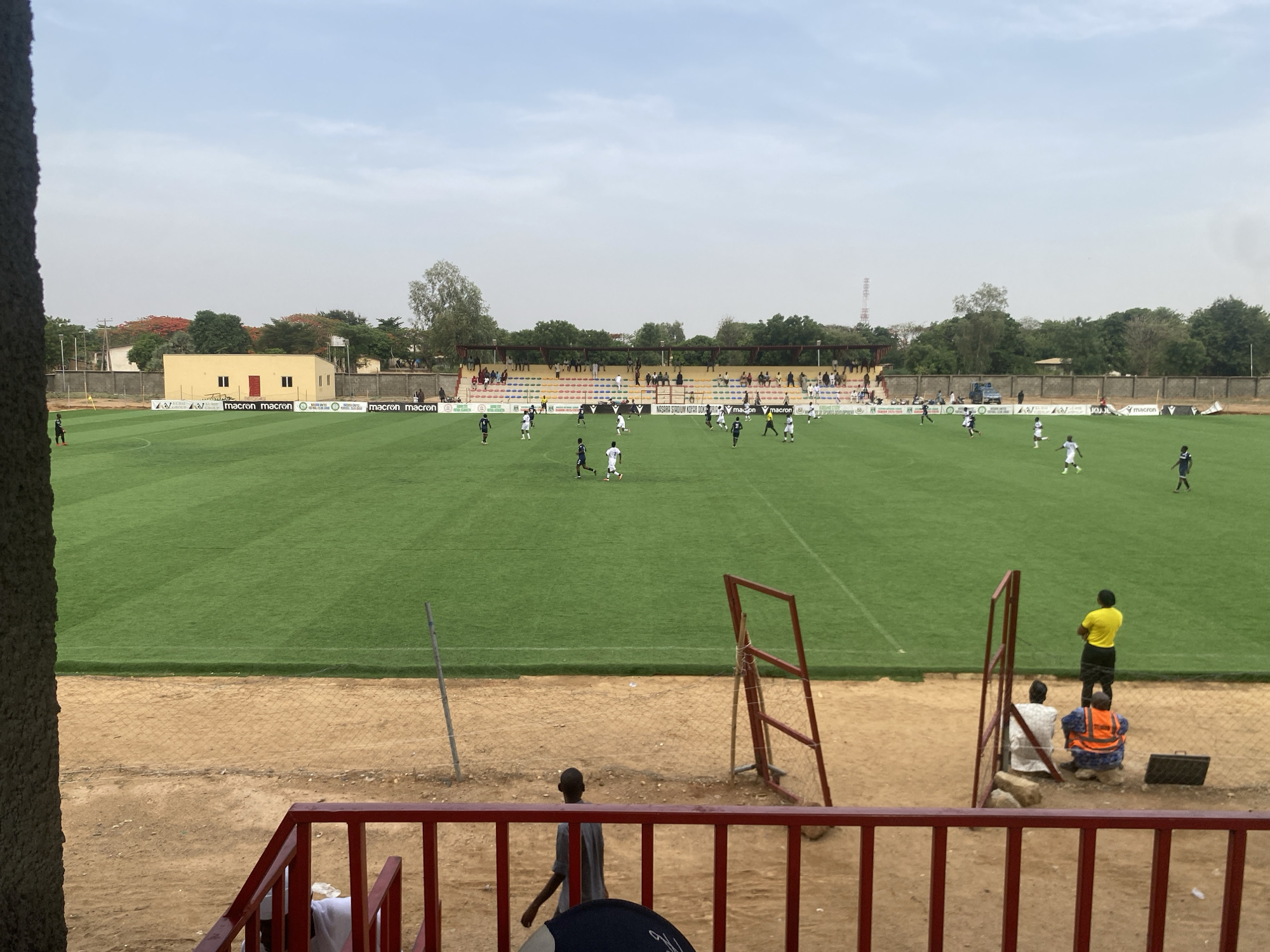 A wide-angle, sunlit outdoor photograph depicts a soccer match in progress at a stadium in Tudun Wada, Nigeria. The well-maintained green field is populated with players wearing predominantly white and dark blue uniforms, engaged in active play. A single-story yellow building stands behind the soccer field, adjacent to an advertising banner bordering the field perimeter. Spectators are seated in a distant stand, which has a small roof. A red railing is visible at the bottom of the frame, along with individuals observing the match from behind a chain-link fence, including a person in a yellow shirt and others in more traditional attire. A structure is partially visible in the left foreground. The sky is overcast, and the overall environment suggests a daytime event.