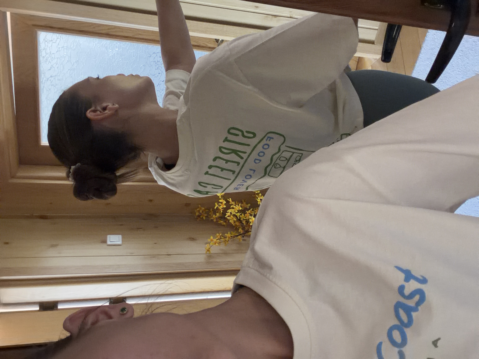 An indoor scene captured from a low angle shows two individuals within a space characterized by wooden paneling. The image, rotated 90 degrees clockwise, depicts a cozy, rustic environment.
In the mid-ground (appearing on the left when viewed upright), a person is visible from the chest up, in profile, looking upwards with their right arm raised. Their dark hair is gathered in a bun. They are wearing a light-colored t-shirt featuring green, reversed text that reads "STREET FOOD LOVER," accompanied by a graphic resembling a food truck or stall. Dark leggings or trousers are visible below their waist.
In the foreground (on the right when viewed upright), a partial view of another person's head and shoulder is seen. Only their right ear and a portion of their hair are clearly visible. They are wearing a light-colored t-shirt with blue text that says "Coast," part of which is cut off by the frame.
The background includes a wooden-framed window with textured, frosted glass, allowing ambient daylight to enter the room. Below the window, on the wooden wall, some yellow artificial flowers or dried foliage are arranged. A white light switch is also visible on the wooden wall. A dark grey cushioned surface, possibly part of a chair, is partially visible in the bottom left. Above the person on the left, a dark brown wooden beam or furniture element crosses the top of the frame.
The activity suggests a casual moment, possibly stretching or reaching for something. The natural light indicates it is daytime. The location context is Buhovo, Bulgaria, although no specific landmarks are visible.