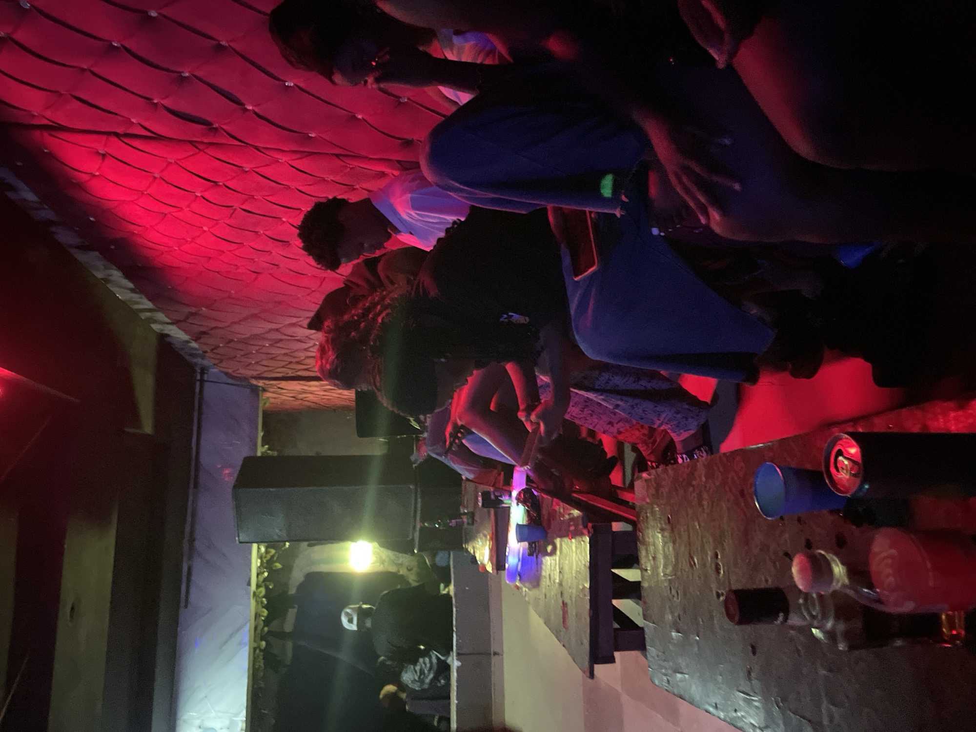 The image depicts a dimly lit indoor scene, likely a club or entertainment venue, illuminated by red lighting and some ambient bluish and yellowish light. Several individuals, appearing to be young adults, are seated on long wooden benches or stools arranged around wooden tables. The people are facing forward, with some engaged in conversation or looking at their phones.
On the tables, various objects are visible, including bottles of alcohol, a can of soda, and a blue plastic cup. The walls of the venue appear to be padded with a tufted design, reflecting the red light. A large black speaker is positioned in the background, and decorative foliage hangs from a ceiling element. The overall atmosphere suggests a social gathering or nightlife setting.
The location specified is the city of Durimi, Nigeria. The scene is consistent with a nightclub or bar environment often found in urban areas.