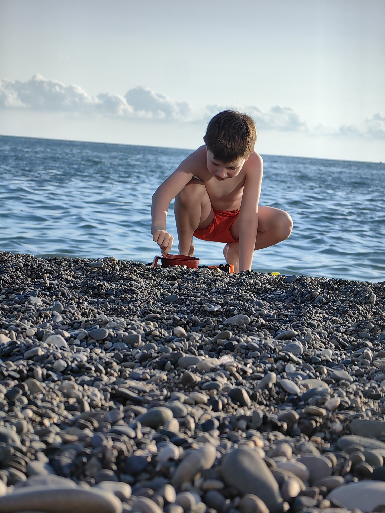 A young boy wearing red swim trunks crouches on a pebbled beach, looking down at a red bucket and toy shovel. The sea is calm and blue behind him, with clouds in the pale sky. The image appears to be taken on a sunny day at a beach in Sochi, Russia.