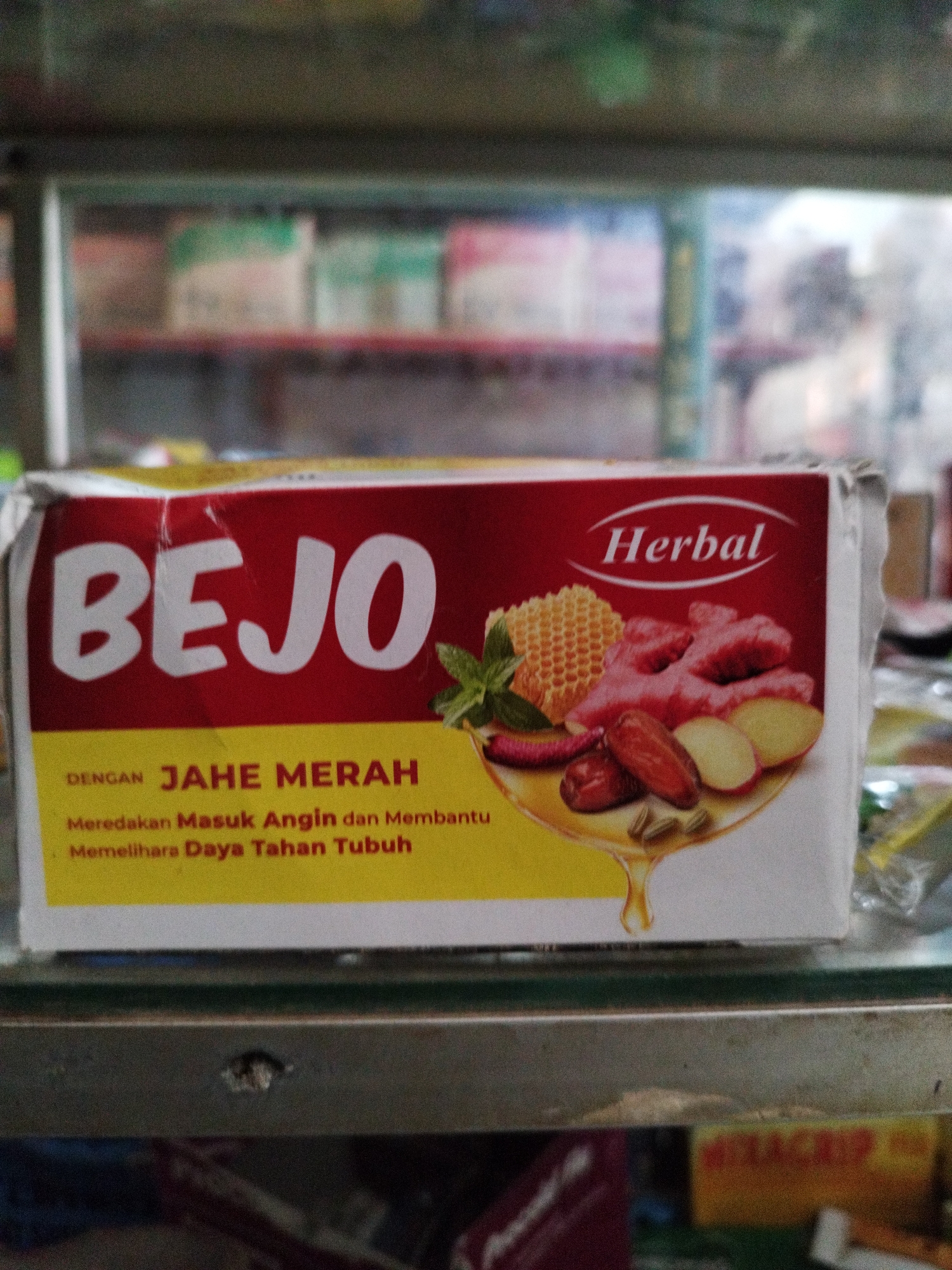 An outdoor, slightly blurry, eye-level, medium shot shows a close-up of a box of "BEJO Herbal" positioned on a shelf in a retail setting. The box is predominantly red with yellow accents and features images of ginger, dates, and herbs. Text on the box indicates it contains "JAHE MERAH" (red ginger) and claims to "Meredakan Masuk Angin dan Membantu Memelihara Daya Tahan Tubuh" (relieve cold symptoms and help maintain the body's immunity). The background displays out-of-focus shelves filled with various products, suggesting it is a small shop or pharmacy. The shelf the box is on appears to be made of metal. The general lighting is dim.