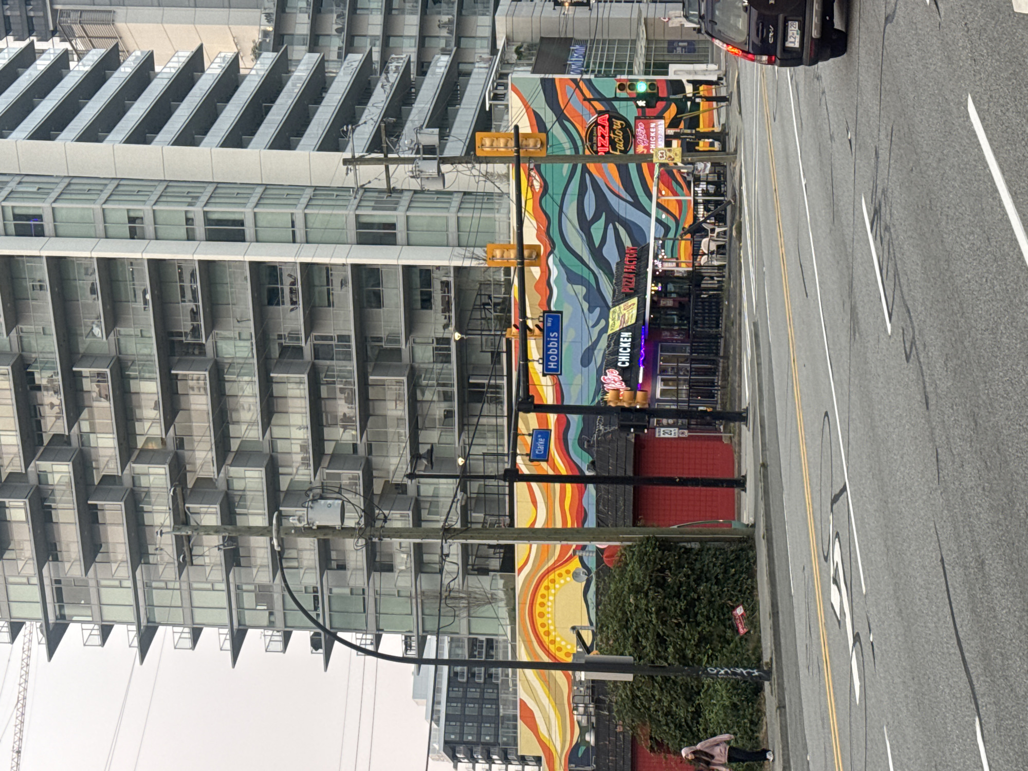 The image depicts a street scene in Coquitlam, Canada, featuring a vibrant mural on the side of a building. A modern, multi-story building with numerous balconies towers behind the mural. In the foreground, a street with lane markings and a bicycle lane is visible. A black SUV is parked on the right side of the road.
The mural itself is characterized by a wavy pattern of bright colors, including shades of orange, yellow, red, blue, and green. It appears to depict a stylized sun or sunrise over a landscape. Below the mural, there are storefronts with neon signs, one reading "CHICKEN" and another "PIZZA FACTORY."
Traffic signals and street signs are present, including one that reads "Clarke" and another "Hobbis Way." Electrical poles and wires are visible, contributing to the urban environment. A person wearing a pink hooded jacket and dark pants is walking on the sidewalk to the left of the frame, with a dense green hedge next to them. The sky is overcast, suggesting a cloudy day. The overall impression is a bustling urban setting with a striking artistic element.