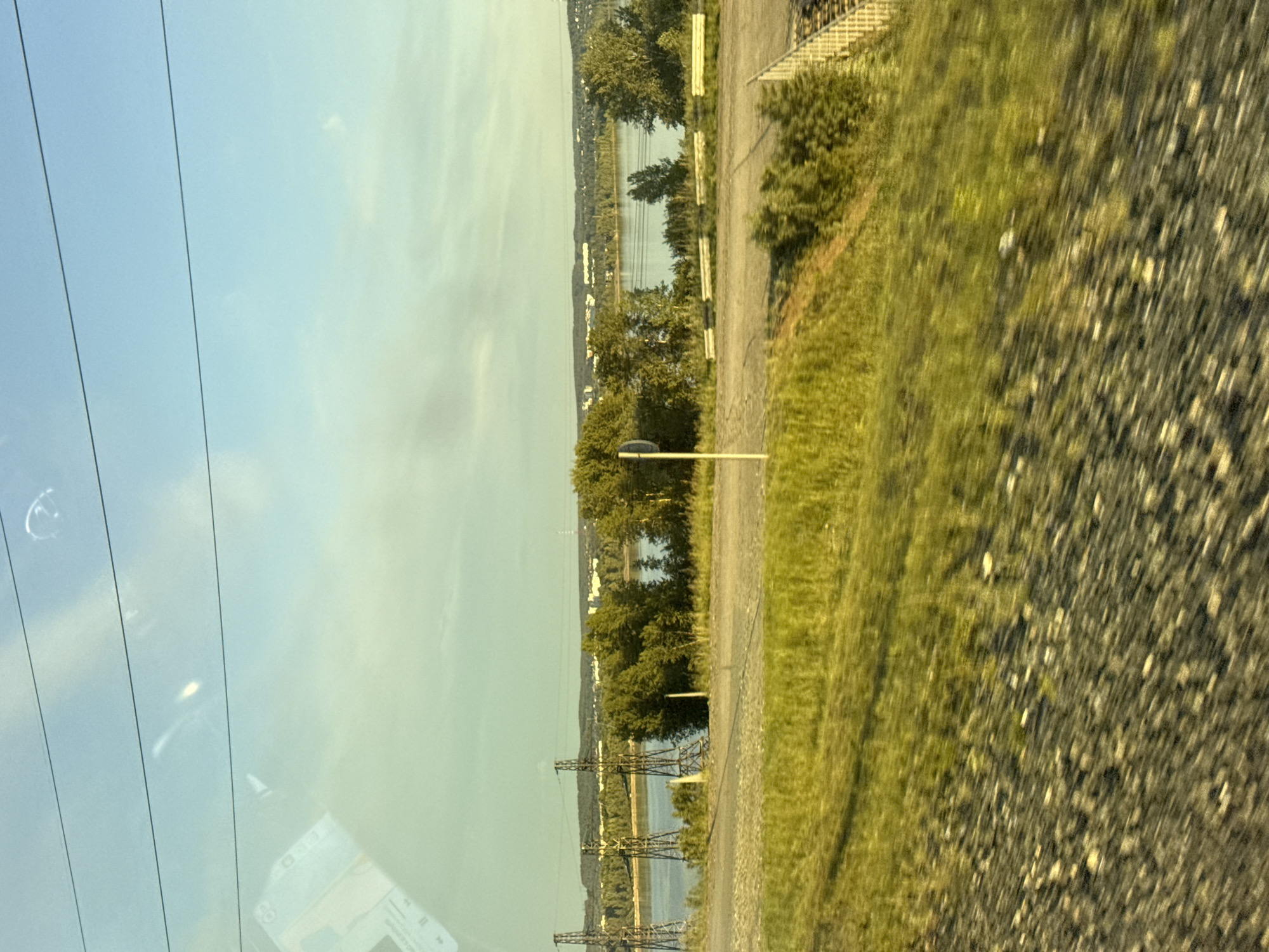 The image displays a landscape scene, likely captured from a moving vehicle, with a distinct horizontal split between the sky and the land/water. The original image is rotated on its side; when viewed upright, the horizon runs horizontally across the mid-section.
In the foreground, on the right side of the image, there is a grassy area transitioning into what appears to be a gravel path or road. This foreground shows some motion blur, particularly on the gravel. On the left side of the foreground, there are significant reflections, suggesting the photo was taken through a window, possibly a car windshield. These reflections include what looks like the interior of a vehicle or a digital display, with faint, indistinct text or symbols, possibly "80" and some graphical icons like play/pause buttons.
The midground features a large body of water, likely a river or a wide lake, reflecting the sky. Trees line the banks of this water body, appearing dense in some sections. Multiple power lines and tall metal power line towers are prominent features, stretching across the landscape and over the water, indicating infrastructure.
In the background, on the far side of the water, a distant landmass is visible, covered in more trees and scattered with what appear to be small buildings or settlements along the shoreline. The sky above is mostly clear, a light blue color on the left, transitioning to a lighter, possibly hazy or slightly yellow hue on the right. The lighting suggests it might be early morning or late afternoon, with a soft, diffused sunlight.
The setting is an outdoor, natural environment interspersed with significant human infrastructure, consistent with a rural or semi-urban area near a large water body, specified as Chaikovsky, Russia. There are no discernible people or explicit activities taking place.