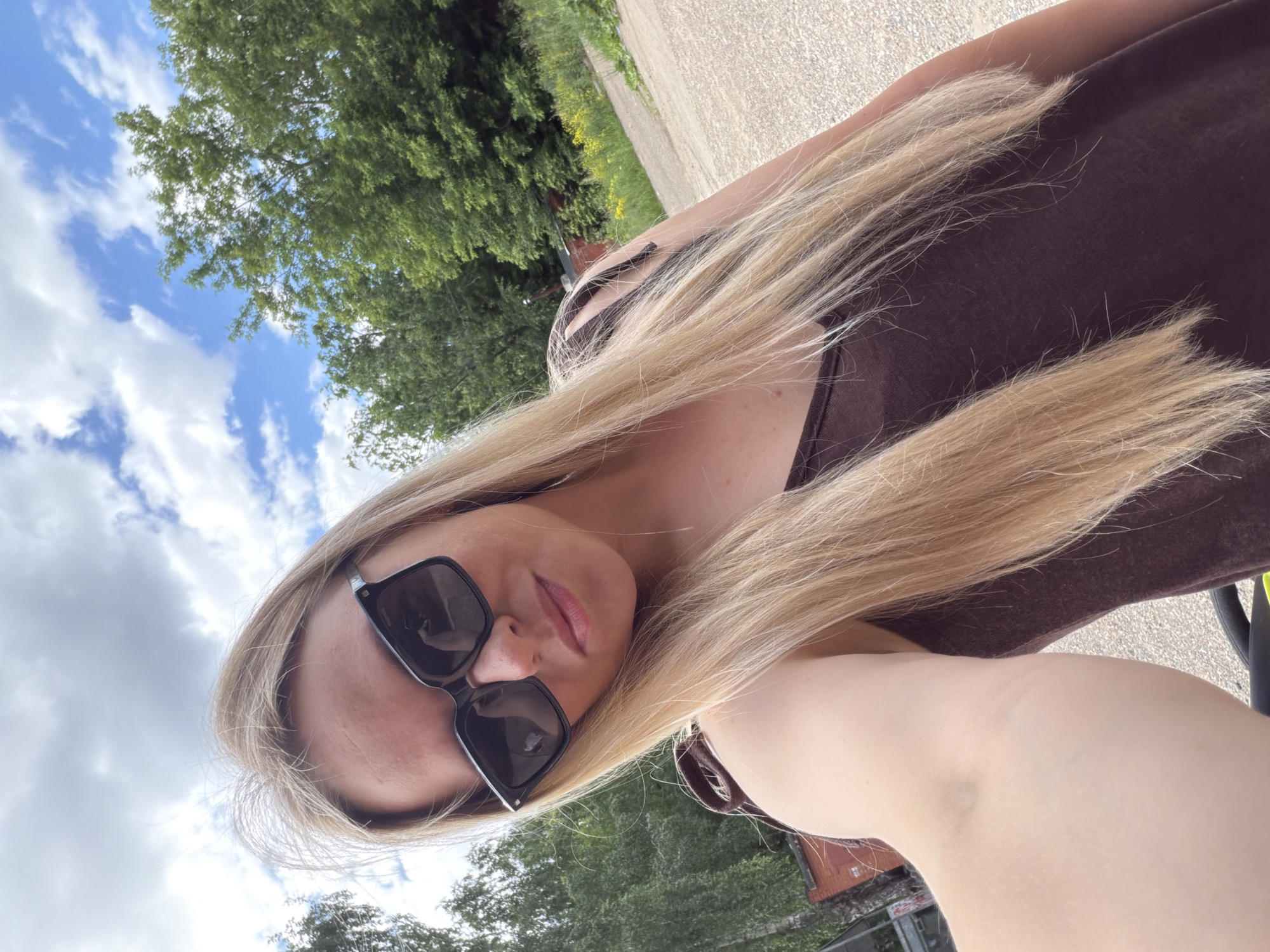 A young woman with long blonde hair, wearing large black sunglasses and a dark brown tank top, takes a selfie outdoors. She has light skin and is looking at the camera with a slight smile. Her hair falls loosely around her shoulders and face. The background is a mix of trees, a cloudy blue sky, and a gravelly path. There are some reddish-brown brick buildings and some yellow flowers visible in the distance. The image appears to have been taken on a bright, sunny day, likely in Votkinsk, Russia, given the context of previous requests.