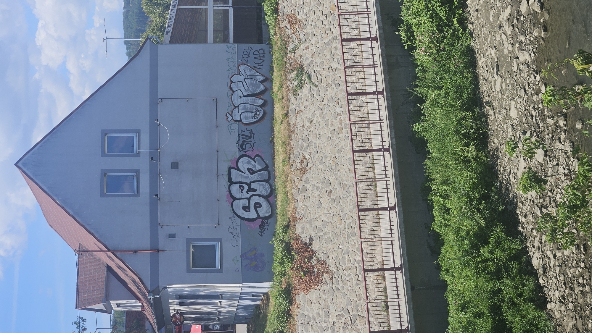 The image shows a building in Prešov, Slovakia, with graffiti on its exterior. The building is a two-story structure with a red tiled roof and light gray walls. Two windows are visible on the upper floor, and a larger rectangular panel is located on the lower floor, possibly a former storefront or advertisement space. Below this panel, graffiti in large bubble letters covers the wall. The graffiti consists of the words "SEK," "ORZ," and "ACAB" along with some smaller tags. The building is situated next to a stone-lined embankment with a red metal railing running along its top. Lush green vegetation grows on the embankment and in the foreground, near a rocky riverbed. The sky above is blue with scattered white clouds. The area appears to be a public space, likely near a residential or commercial district.