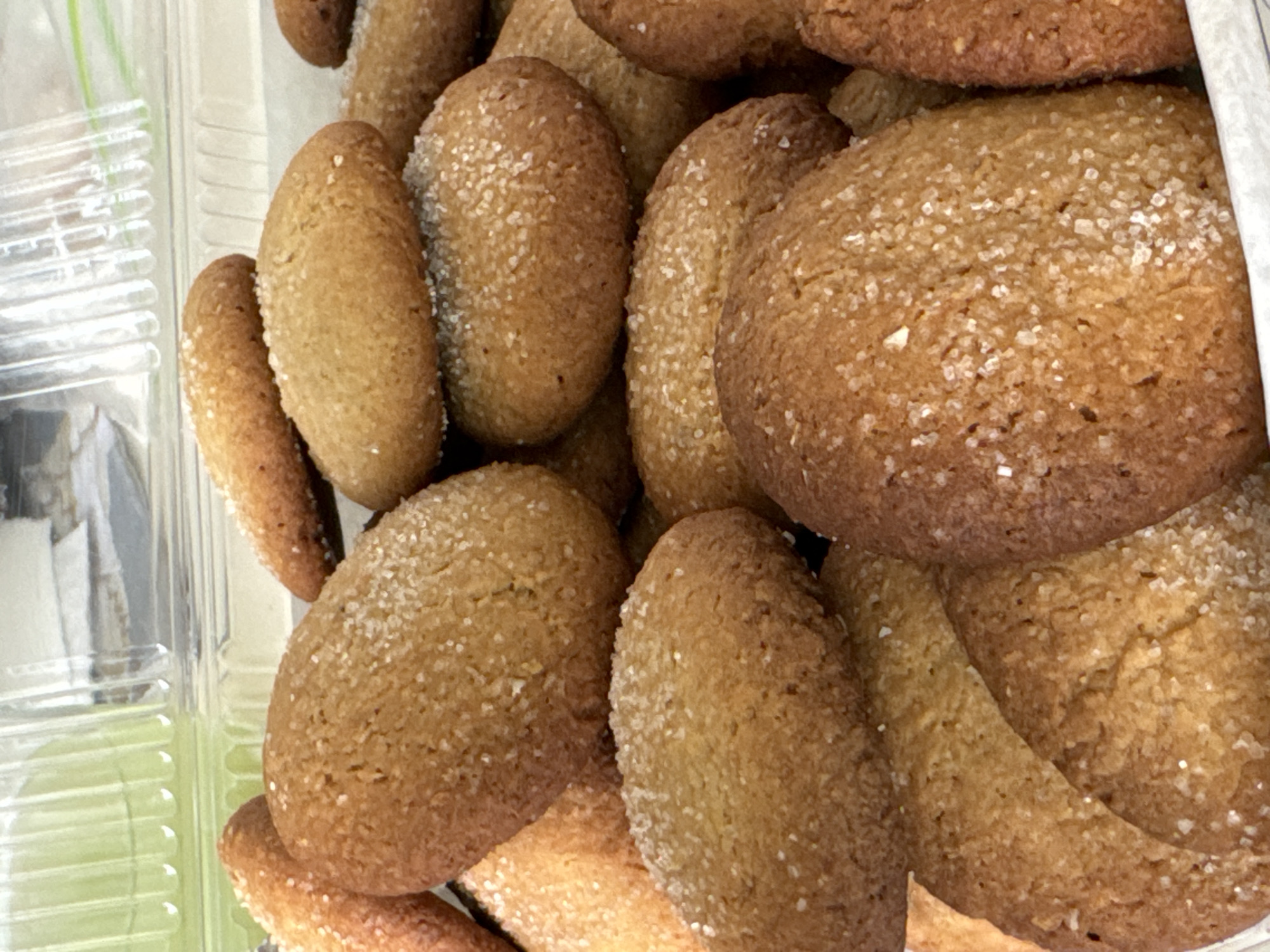 A close-up shot shows a pile of brown sugar cookies, some oval-shaped and some round, densely packed in a clear plastic container. The cookies are textured and covered with coarse sugar crystals. Part of the plastic container is visible on the left side, with a green surface possibly behind it. A white object partially covers the cookies at the upper right. The scene is indoors, given the context of a container and likely artificial lighting, but the specific location in San Bernardino Tlaxcalancingo, Mexico, is not inferable from the visual data.