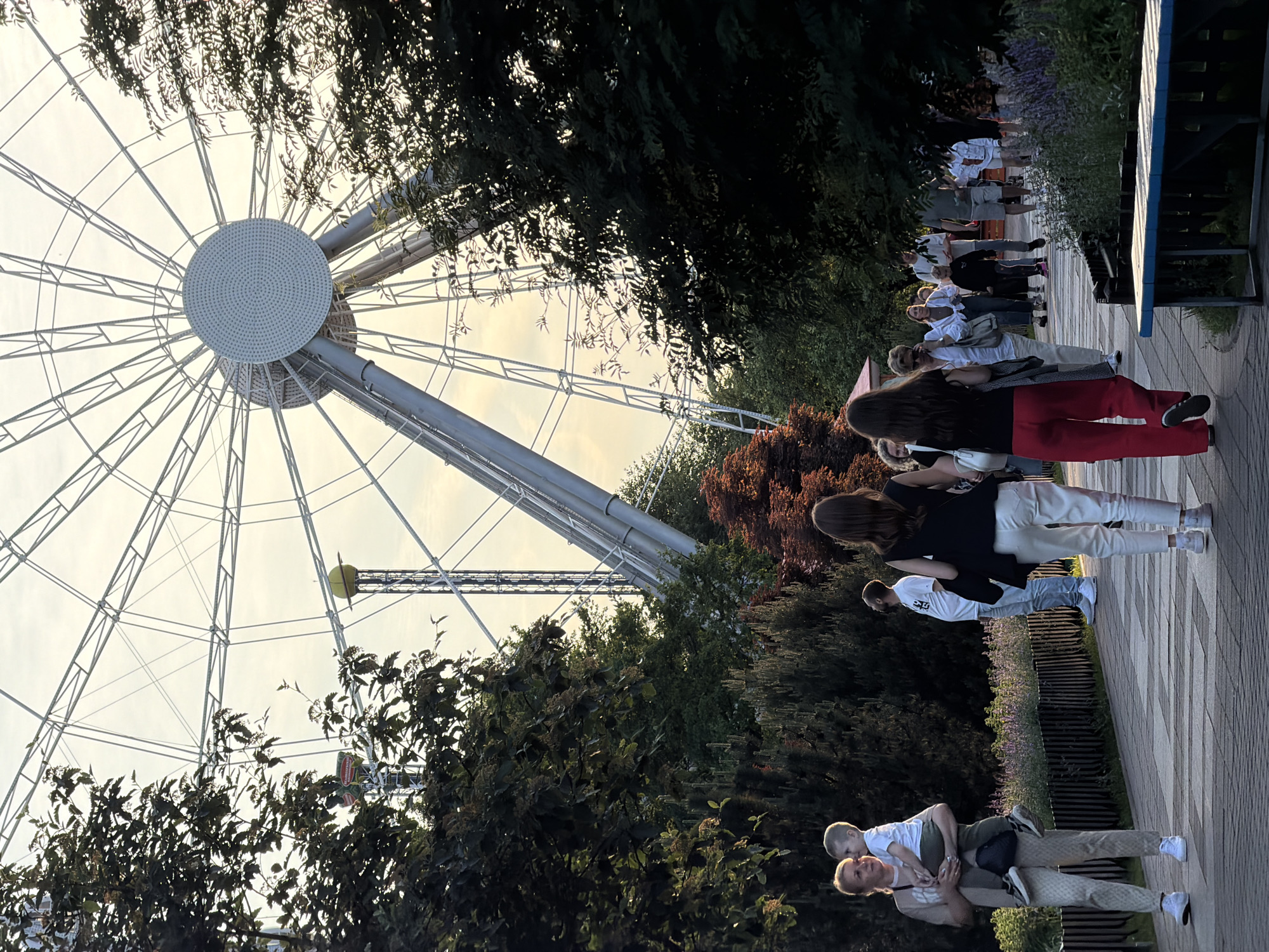 The image depicts a Ferris wheel at the "Divo Ostrov" amusement park, located on Krestovsky Island in Saint Petersburg, Russia. The Ferris wheel dominates the background, its large white structure with radiating spokes reaching towards a pale sky. In the foreground, people are walking along a paved pathway lined with trees and flowering bushes.
On the left side of the pathway, a woman is holding a young child. She is wearing a light-colored top and patterned trousers, with white sneakers. The child, dressed in a white t-shirt and dark pants, is looking towards the woman.
Further along the path, a woman in a black top and white trousers walks away from the camera. To her right, a man in a white t-shirt and light blue jeans stands facing away, looking upwards. More people are visible in the middle ground and background, some walking and others seemingly gathered.
The environment suggests a pleasant outdoor setting, likely during daytime or early evening, given the lighting. The presence of the Ferris wheel and the pathways indicates a public recreational area. The trees are lush and green, with some foliage showing autumn colors in the middle ground.