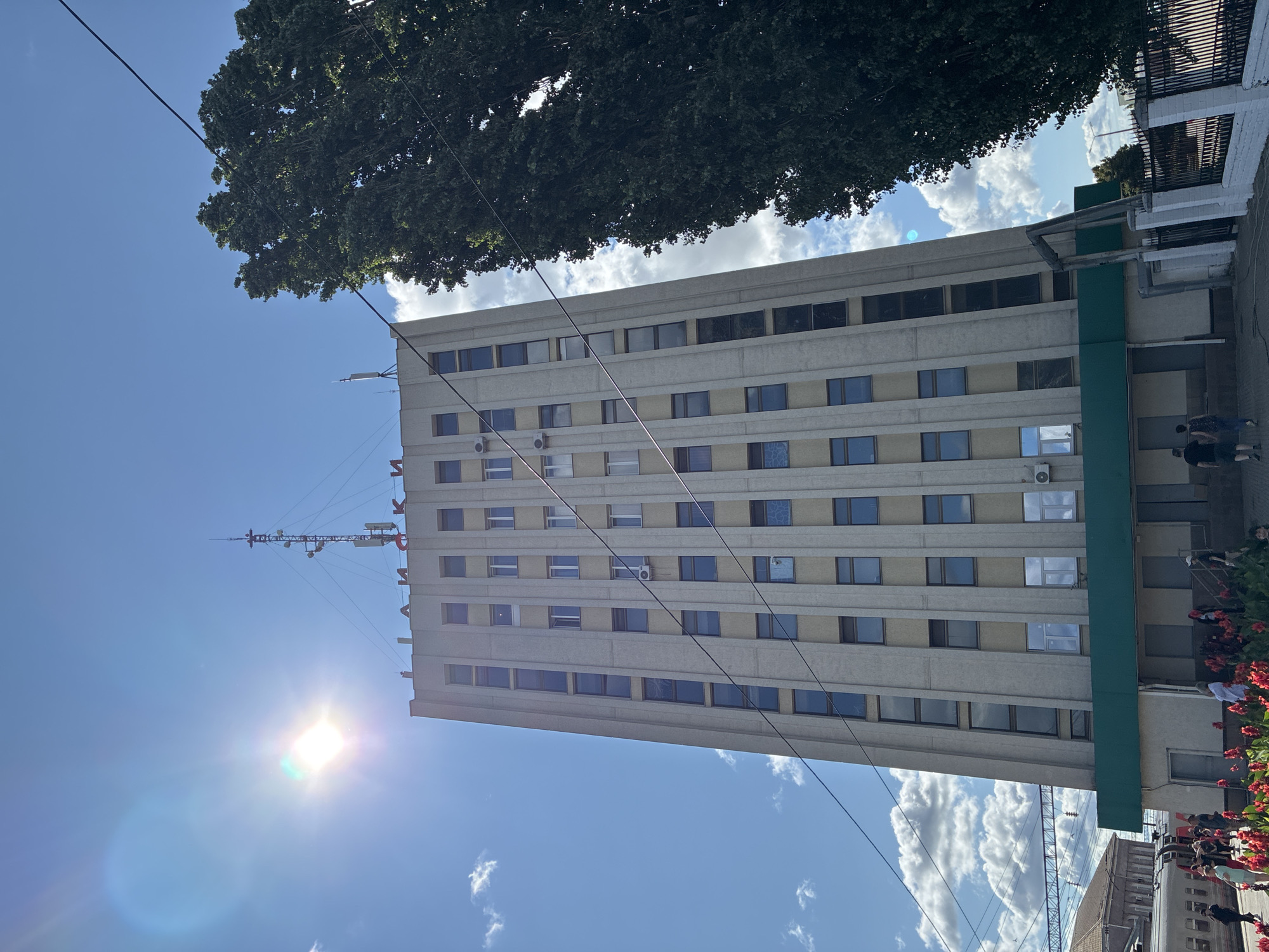 The image shows a tall, rectangular administrative building with a light-colored facade and vertical window panes. Red Cyrillic lettering is visible at the top of the building, spelling out "ЛИСКИ" (Liski). The building has a green awning at its base. To the right of the building, a tall, dark green tree with dense foliage dominates the frame. The sky is bright blue with scattered white clouds, and the sun is prominently visible in the upper left corner, casting lens flare. In the foreground, several people are walking and standing, with red flowers in bloom. A train is visible to the left of the building, and railway tracks are also present. The scene appears to be a public area, possibly a train station or a central square in the city of Liski, Russia.
