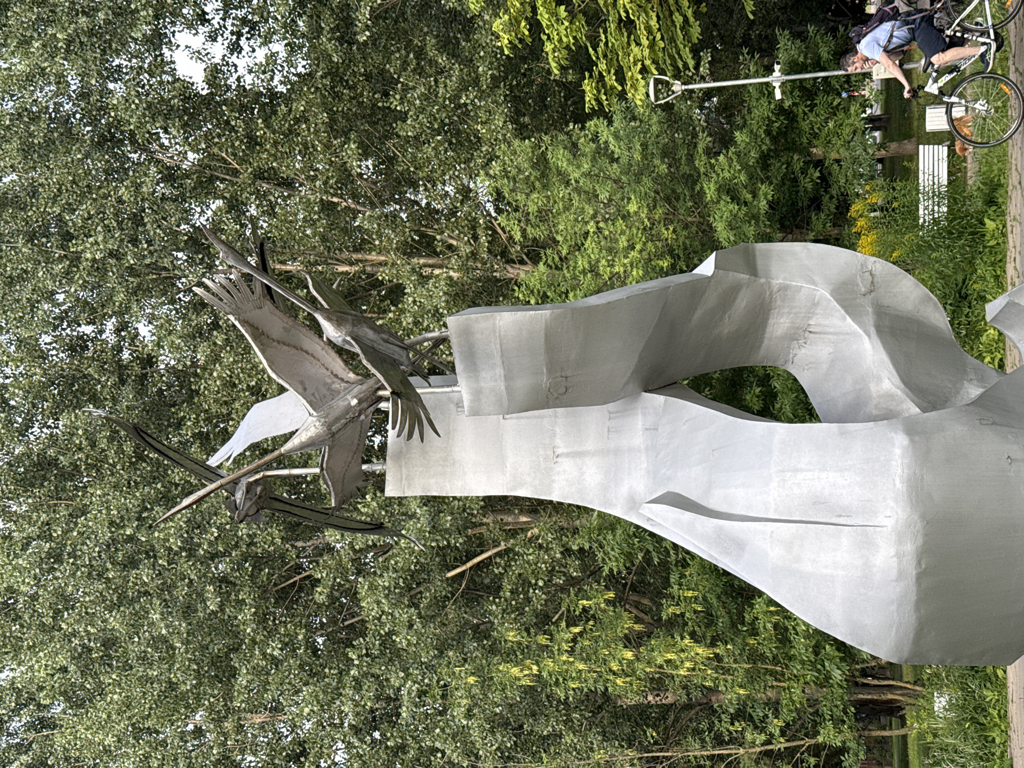 The image captures a kinetic sculpture of birds in flight, likely cranes, made of brushed metal. The sculpture is situated outdoors, with a backdrop of lush green trees and foliage. In the middle ground, a man is riding a bicycle on a paved path, with a light pole and security cameras visible in the background. The overall impression is that of a public park or urban green space. The sculpture appears to be a prominent landmark in a city setting. The sculpture is identified as the Monument to the Fallen Cranes in Moscow, Russia.