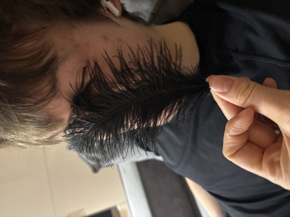The image captures a close-up, rotated view of a person's head and shoulder, with a black feather being held to their face. The person has light brown or blonde hair, which is styled to fall across their forehead and temples. They appear to have their eyes closed or nearly closed, and there are several small blemishes or acne spots visible on their cheek and forehead. A white wireless earbud is visible in their right ear. They are wearing a plain black t-shirt or top.
A hand, presumably belonging to another person, is holding a long, black, feathery plume. The hand has manicured nails with a light, natural-looking polish that has clear tips. The thumb and index finger are pinching the stem of the feather, positioning the soft, feathery part against the first person's face, specifically near their cheek and eye. The act appears to be a gentle tickle or a playful gesture, eliciting the closed-eye reaction.
The background is softly blurred, suggesting an indoor setting. Muted, light brown and grey tones are visible, possibly indicating furniture like a couch or a wall. The overall lighting is soft, likely ambient indoor light. The image's rotation suggests it might have been taken with the phone held at an angle or that the subject was lying down. There is no visible text in the image.