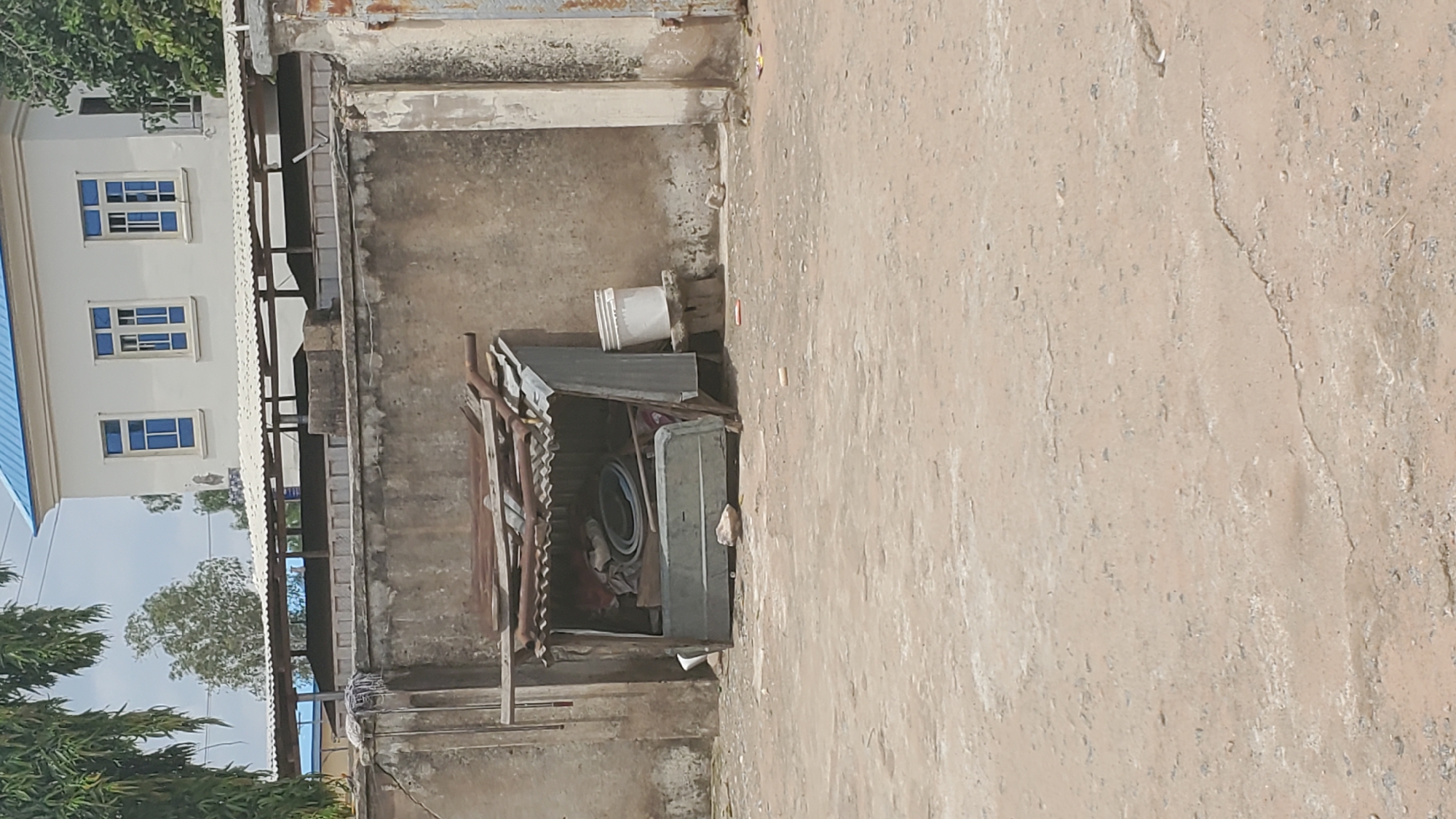 This is a vertical, medium-distance shot of an outdoor scene in Jalingo, Nigeria. In the foreground is a dusty, uneven ground. In the middle ground, against a rough, weathered wall, is a makeshift shelter constructed from corrugated metal and wood scraps. The shelter is open on one side, revealing various items stored inside, including what appears to be folded fabric and plastic containers. To the right of the shelter, a white bucket sits on a low, stone-like structure. Behind the shelter and wall, a large building with a bright blue roof and white walls is visible, featuring multiple windows with blue frames. Trees are present in the background, with one prominently positioned to the left, its green foliage casting shadows. The sky is overcast.