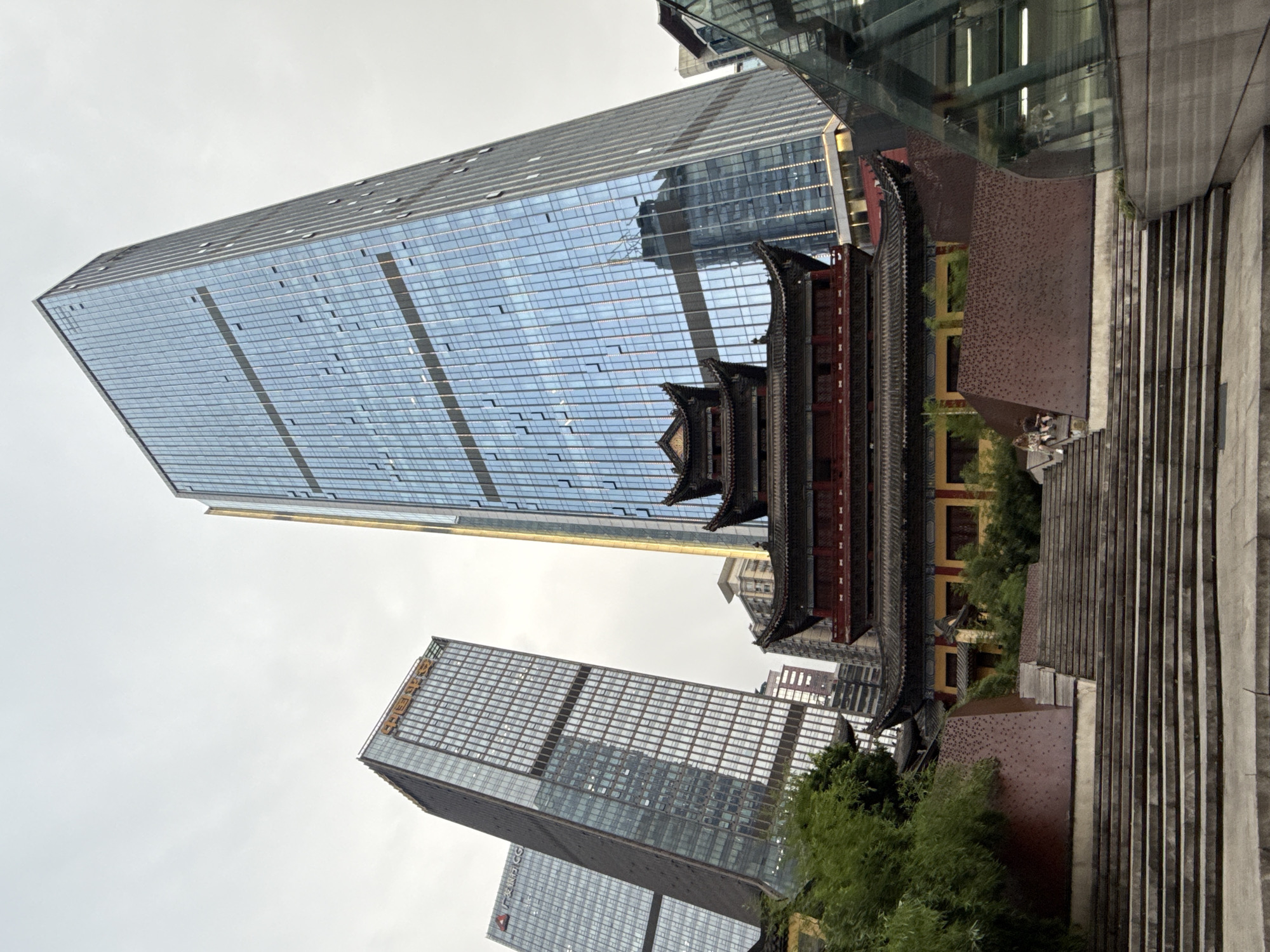 The image, taken from a low angle in Yuzhong District, China, captures a striking juxtaposition of traditional architecture and modern skyscrapers. In the foreground, a wide, tiered stone staircase ascends towards a traditional Chinese temple with dark, tiered roofs and red accents. Behind the temple, two imposing, modern glass skyscrapers rise into the overcast sky. The skyscraper on the right has a vertical golden stripe running up its facade, while the one on the left, slightly further back, displays Chinese characters in orange near its top. Another skyscraper, partially visible on the far left, also features a logo. The scene is populated by a few individuals, including two people sitting on the steps of the temple area, and a reflection of a building in the glass facade of the skyscraper. Lush green trees are visible to the left of the temple, and a modern glass structure partially obscures the right side of the frame. The overall impression is one of an urban landscape where historical and contemporary elements coexist.