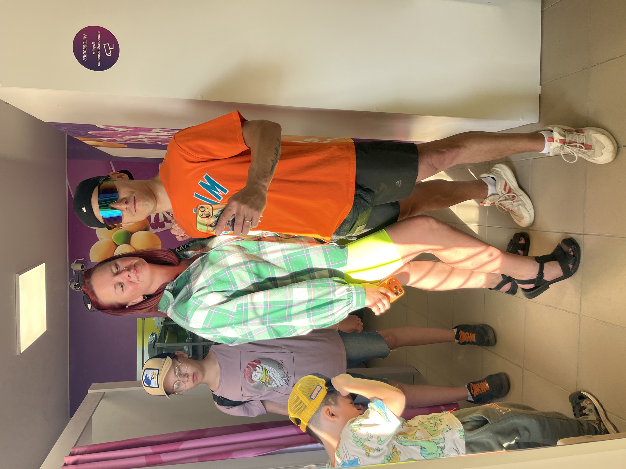 Four people are standing in what appears to be a dressing room or fitting booth. From top to bottom, there is a man wearing an orange t-shirt, black shorts, and sports shoes with white socks. He has a pair of sunglasses on top of his head. Below him is a woman in a green plaid shirt, holding a phone. Under the woman is a teenager wearing a t-shirt with an image on it, denim shorts, and sneakers. The last person is a young boy in a yellow hat, t-shirt with dinosaurs and wearing sweatpants with sneakers. The scene appears to be in Chaikovsky, Russia, based on the signage visible in the background.