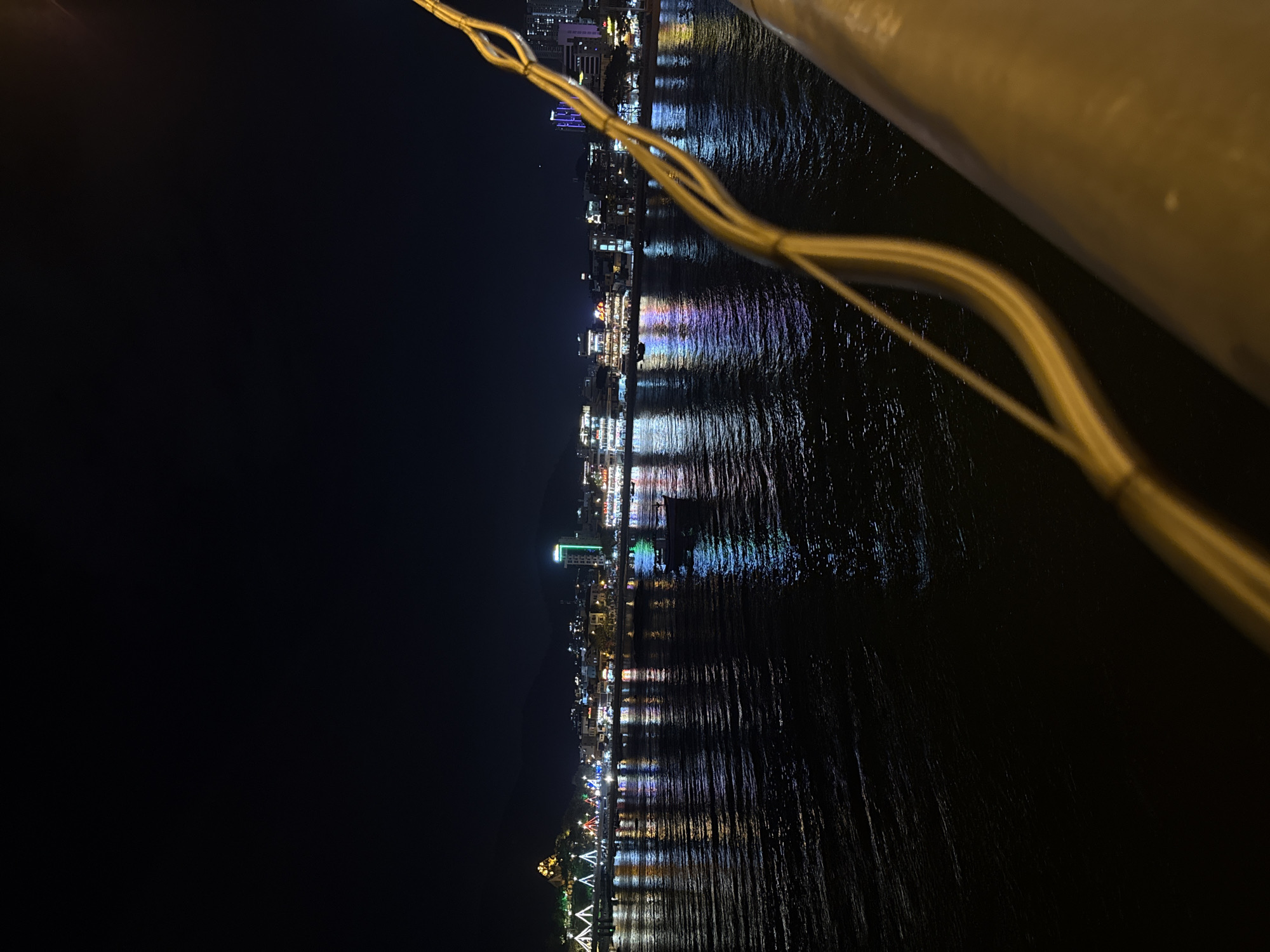 This image captures a nighttime view of the Nha Trang coastline in Vietnam. The foreground is dominated by a blurry yellow railing and some electrical cables, suggesting the viewpoint is from a bridge or elevated structure. In the mid-ground, a dark body of water, likely the sea, reflects the colorful lights of the city skyline. Several boats are visible on the water, their lights adding to the reflections. The city itself is illuminated with a variety of lights, including a prominent green lit building on the right, and a cluster of buildings with colorful lights on the left. A hill with a structure on top, possibly a statue or monument, is visible in the far left of the frame. The sky is dark, indicating it is nighttime.
