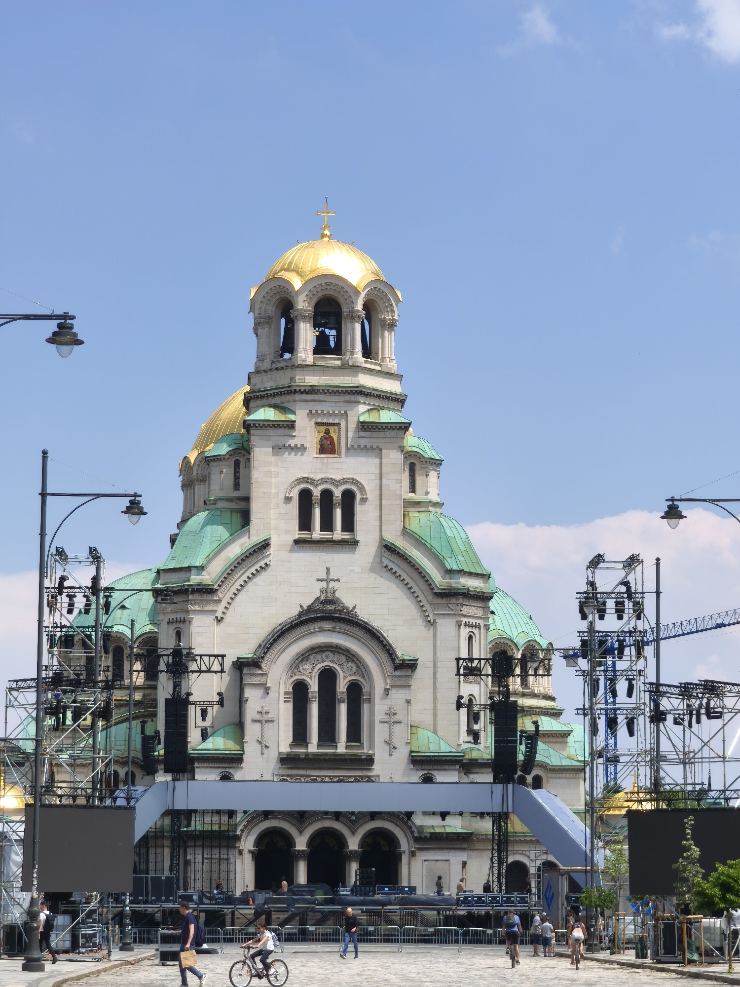 The image depicts the Alexander Nevsky Cathedral in Sofia, Bulgaria, partially obstructed by extensive temporary event infrastructure. The cathedral features white stone walls, prominent golden domes, and verdigris-green smaller domes, all topped with Orthodox crosses. A bell tower is visible on the main golden dome, and an icon is integrated into an upper facade.
In the foreground and midground, a complex network of metal scaffolding, trusses, and staging elements has been erected. This infrastructure includes large speaker arrays (line arrays) suspended from trusses, numerous stage lights, and two large, dark rectangular display screens (likely LED screens) positioned in the lower left and lower right foregrounds. A blue-gray ramp or walkway structure ascends towards a raised stage area that further obscures the lower part of the cathedral. Metal crowd control barriers delineate pathways on the cobblestone-paved ground.
Several individuals are present in the scene. In the foreground, a person walks right-to-left carrying a paper bag, while another person walks left-to-right. Two individuals are cycling: one in the midground on a bicycle, and another further back. Other people are visible walking behind the barriers and on the cobblestone area. Some figures appear to be working on or around the temporary structures.
The sky is clear blue with minimal scattered white clouds. Two black streetlights with circular luminaires are positioned in the left foreground and midground. The overall scene suggests an outdoor public event being set up or in progress in front of the historic cathedral.