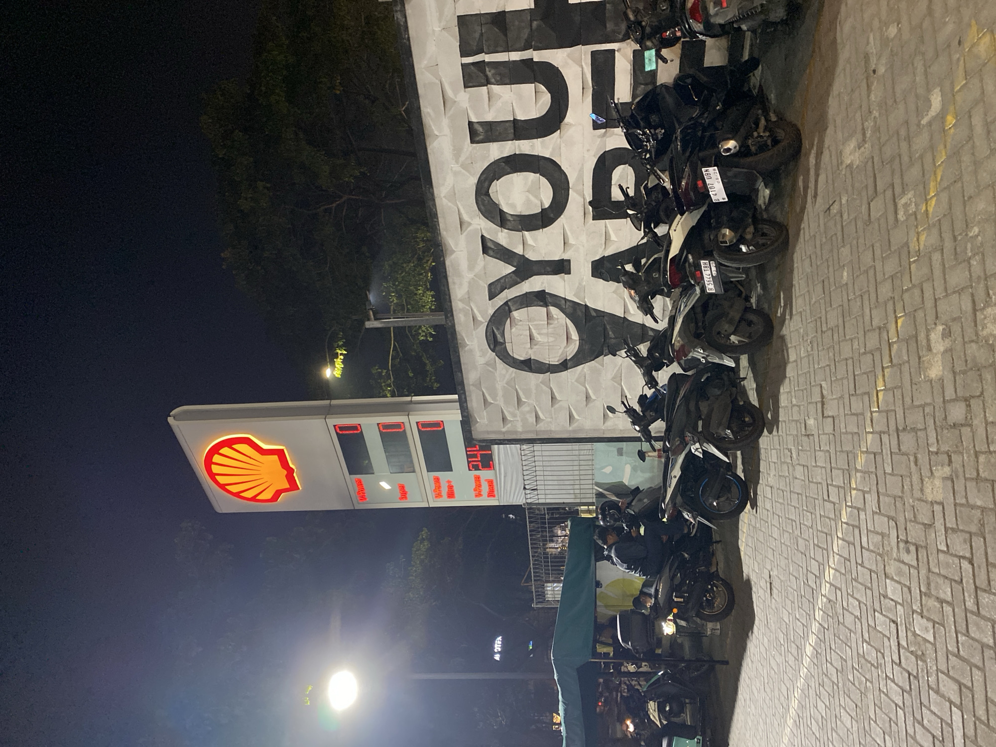 This media file depicts a night scene at a Shell gas station in East Jakarta, Indonesia. The primary subjects are a row of parked motorcycles and a large, illuminated Shell logo sign. The sign features the iconic red and yellow shell emblem. Adjacent to it, a gas pump display shows prices for various fuel types: "V-Power", "Super", "V-Power Nitro+", and "Diesel", with numerical values displayed in red digital characters.
The background is dominated by a textured, white wall with large, black lettering that appears to spell out "OYOUL ADE", though the full phrase is not entirely visible. The ground is paved with light-colored bricks, and yellow lines mark parking or traffic lanes. Several people are visible, mostly seated on or standing near the motorcycles, suggesting they are either waiting or have just arrived. The lighting suggests it is nighttime, possibly late evening or early morning, with artificial lights from the gas station and streetlights illuminating the scene. The atmosphere appears to be calm, with no indication of any urgent activity. The presence of multiple motorcycles and the gas station setting point to a common stop for commuters or travelers. The weather is not explicitly indicated but appears clear.