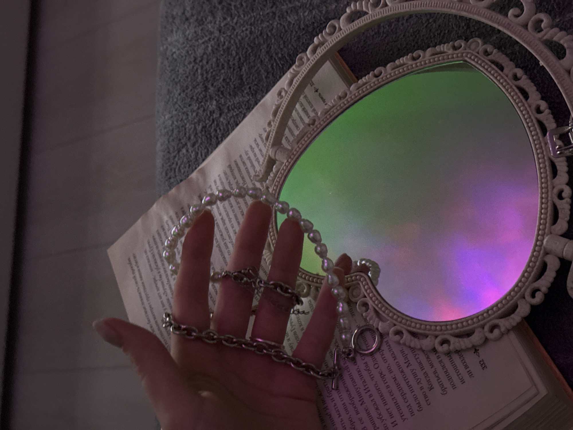 A hand with two chain rings and a pearl necklace lies in front of an open book, which is behind a heart-shaped ornate mirror. The mirror's surface reflects an iridescent green and purple hue. The scene is bathed in a muted, ambient light, casting a soft glow over the objects. The book's open pages reveal text in a Cyrillic script, and the pearl necklace drapes across the hand and the book's edge. The background includes a gray textured surface, possibly a sofa or blanket. The overall composition creates a dreamy, artistic tableau.