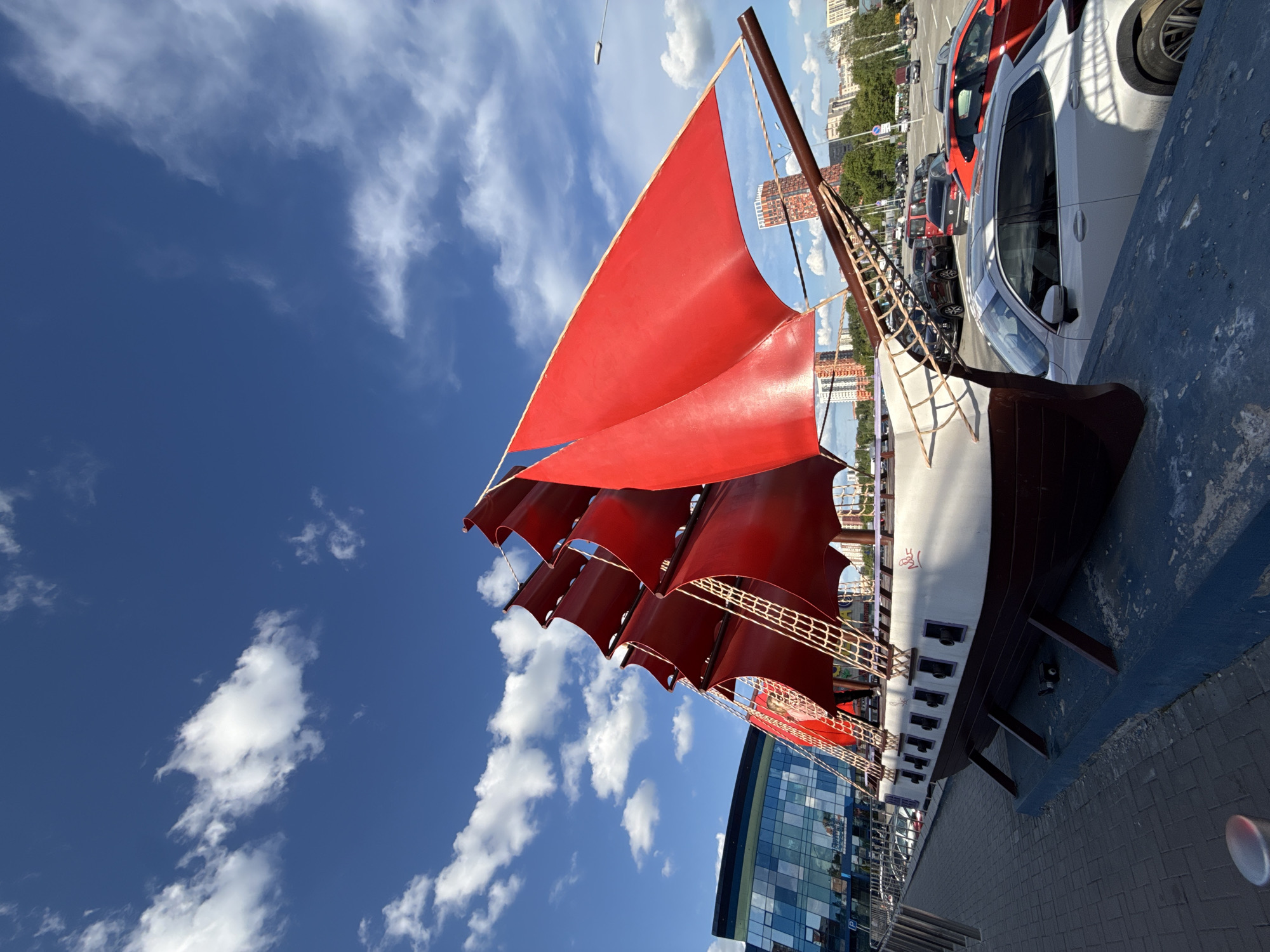 This media file displays a large, elaborate model of a sailing ship under a bright blue sky with scattered white cumulus clouds. The ship, which appears to be a land-based installation, features striking, multiple red sails fully unfurled, supported by dark wooden masts and intricate rigging with ropes and netting. Its hull is designed with a dark brown bottom and a white upper section, complete with several small dark, rounded features resembling portholes or cannons along its side.
In the background, an urban setting unfolds, showing a paved street or parking area densely lined with parked cars of various models and colors, including white, red, and dark vehicles. Beyond the street, there are several multi-story buildings, one of which is a modern structure with reflective glass panels, and others appearing to be residential or commercial buildings. The sunlight is strong, indicating it is daytime with good weather. A small, red, stylized symbol or character is visible on the white part of the ship's hull. The scene is static, with no discernible human activity. The overall impression is of a vibrant, sunny day in Izhevsk, Russia.