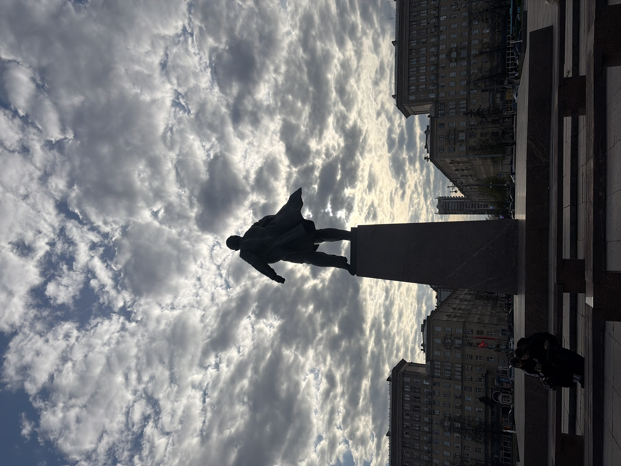 The image shows a statue of Vladimir Lenin in Saint Petersburg, Russia, viewed from a low angle. The statue is silhouetted against a cloudy sky. Buildings are visible in the background, lining a street. A group of two people, who are obscured by shadow, stand near the edge of the platform on which the photographer is standing.